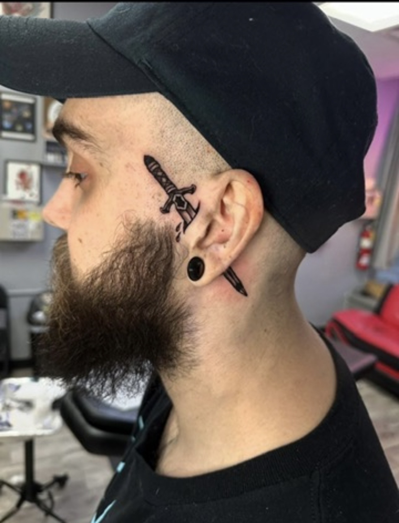Side profile of a man with a beard, wearing a black cap, showing tattooed sword and dagger on his neck.