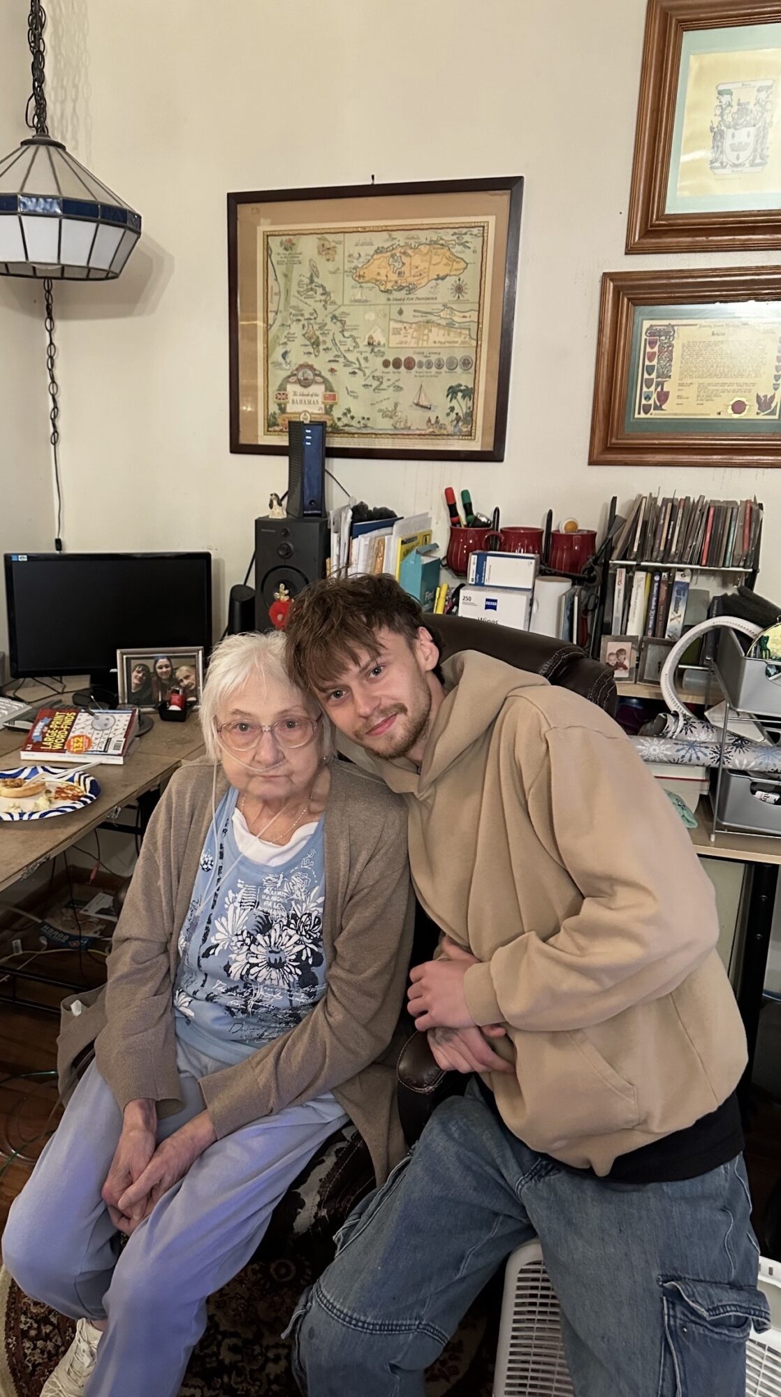 Two people sitting and standing in a room with framed pictures, a computer, and books on shelves behind them.