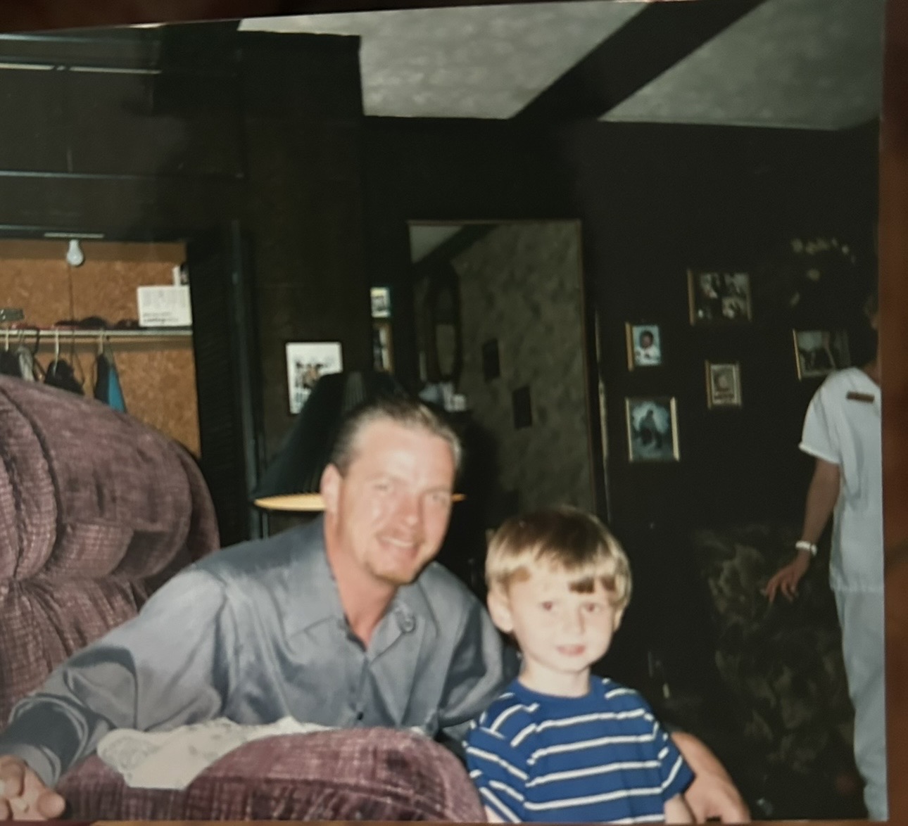A man and a boy sit together indoors, smiling at the camera, with a dark interior background.
