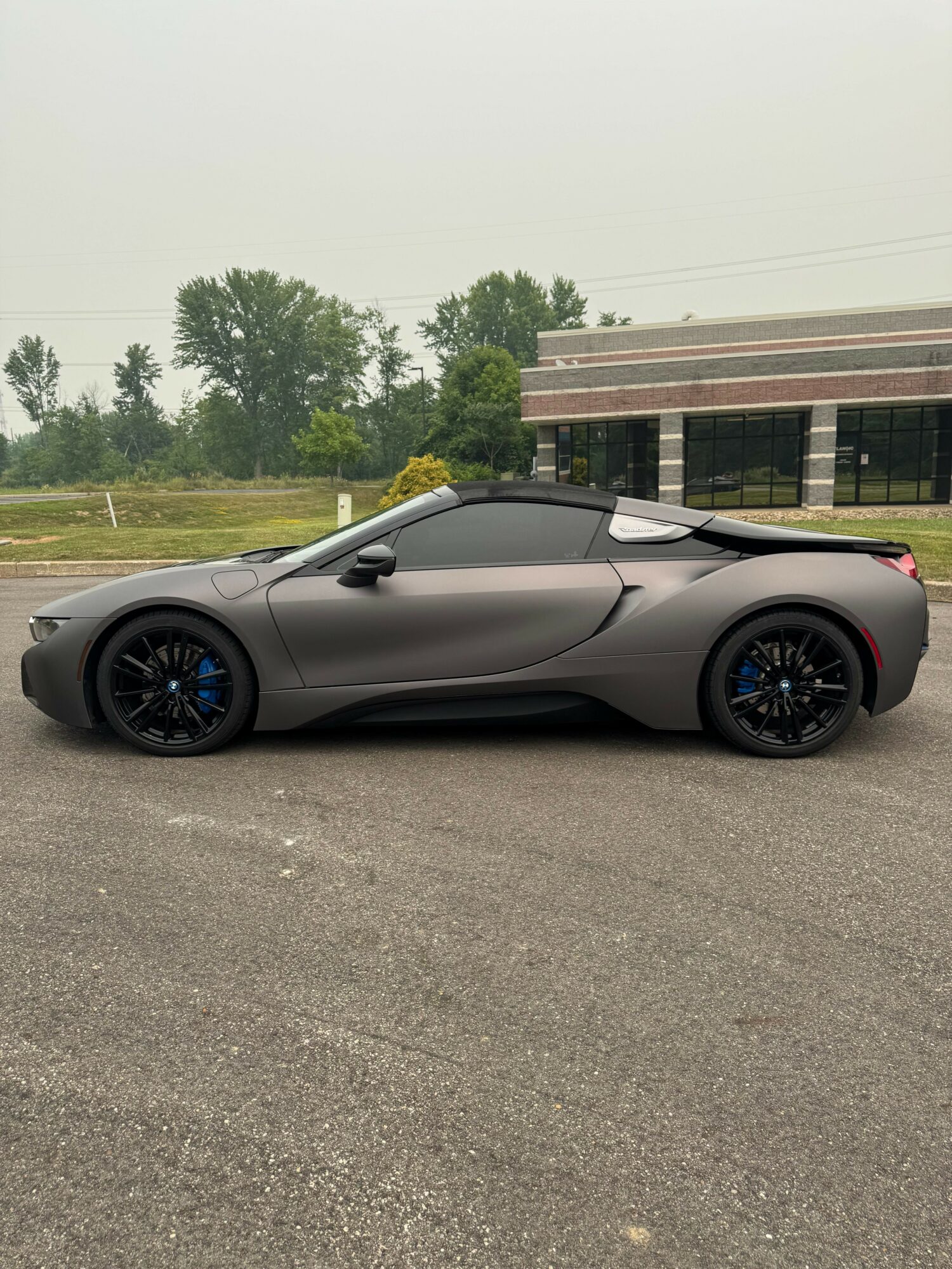 Gray sports car with black and blue wheels parked on a paved surface, with trees and a building in the background.