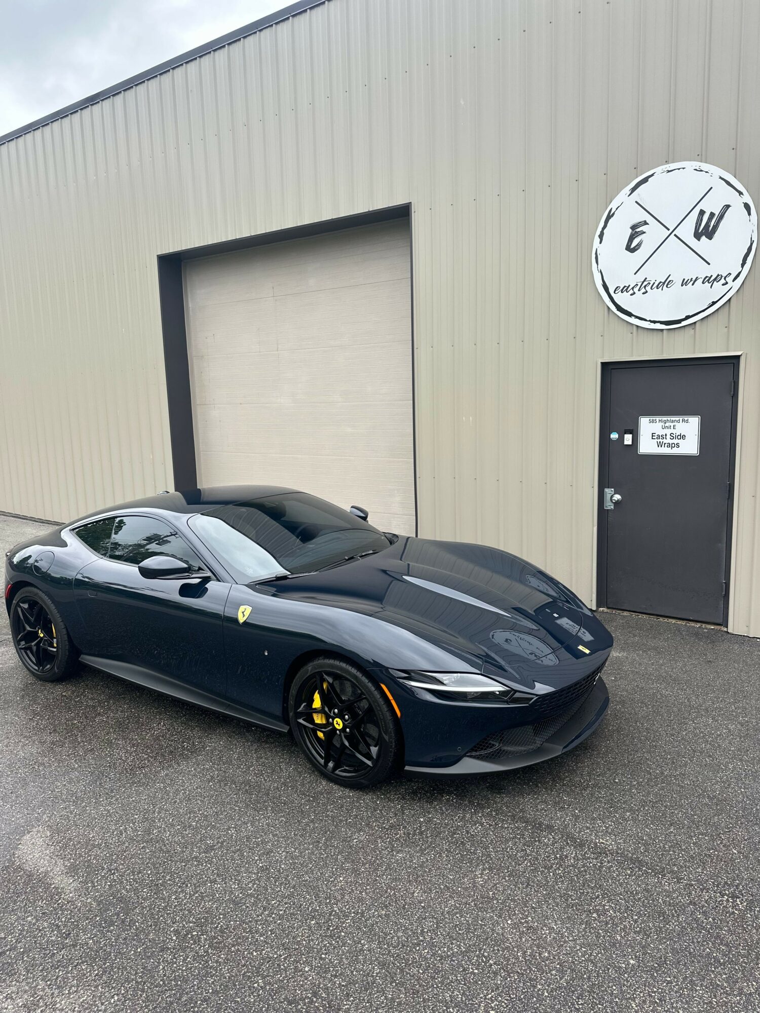 Black sports car parked on gray asphalt in front of a beige building with a large clock and a black door