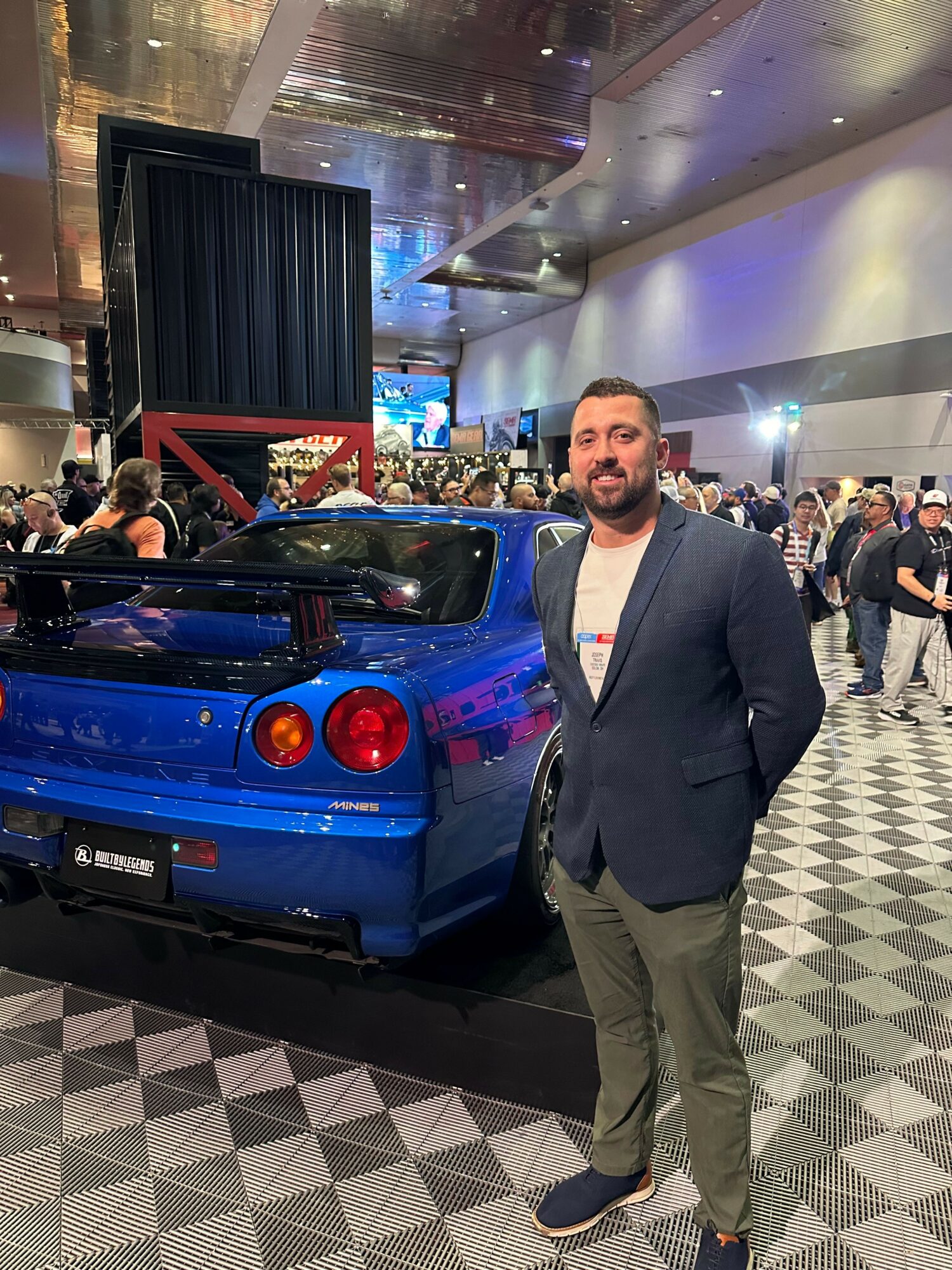 Man in suit standing next to a blue sports car on display in a busy indoor venue.