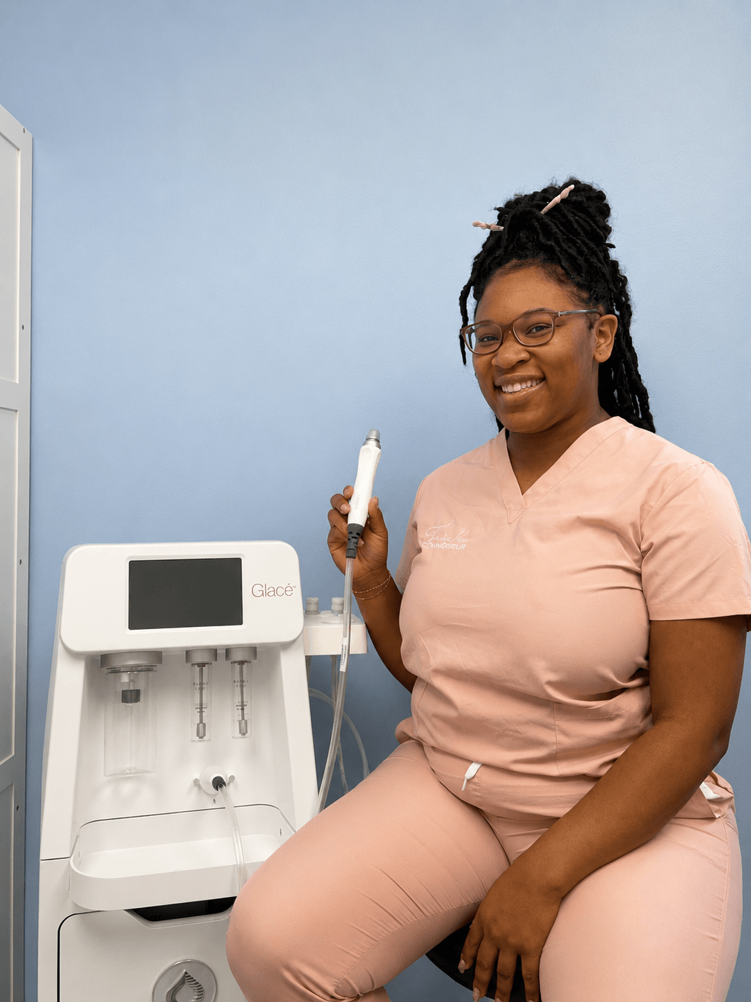 A woman in pink scrubs smiling and holding a medical device, standing next to a medical machine against a blue wall.