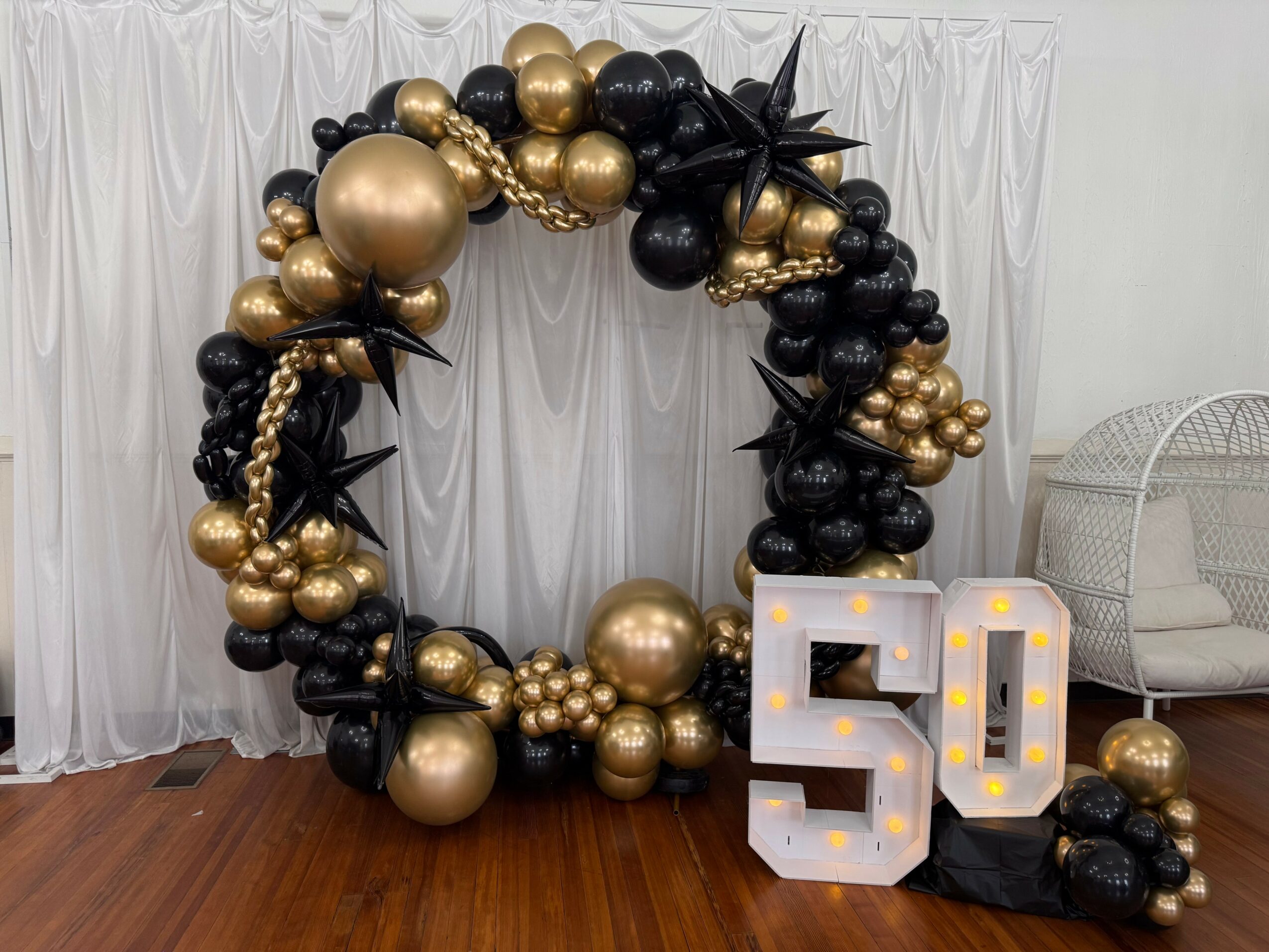 Decorative balloon arch with black and gold balloons, large illuminated number 50, and a white chair in the background.