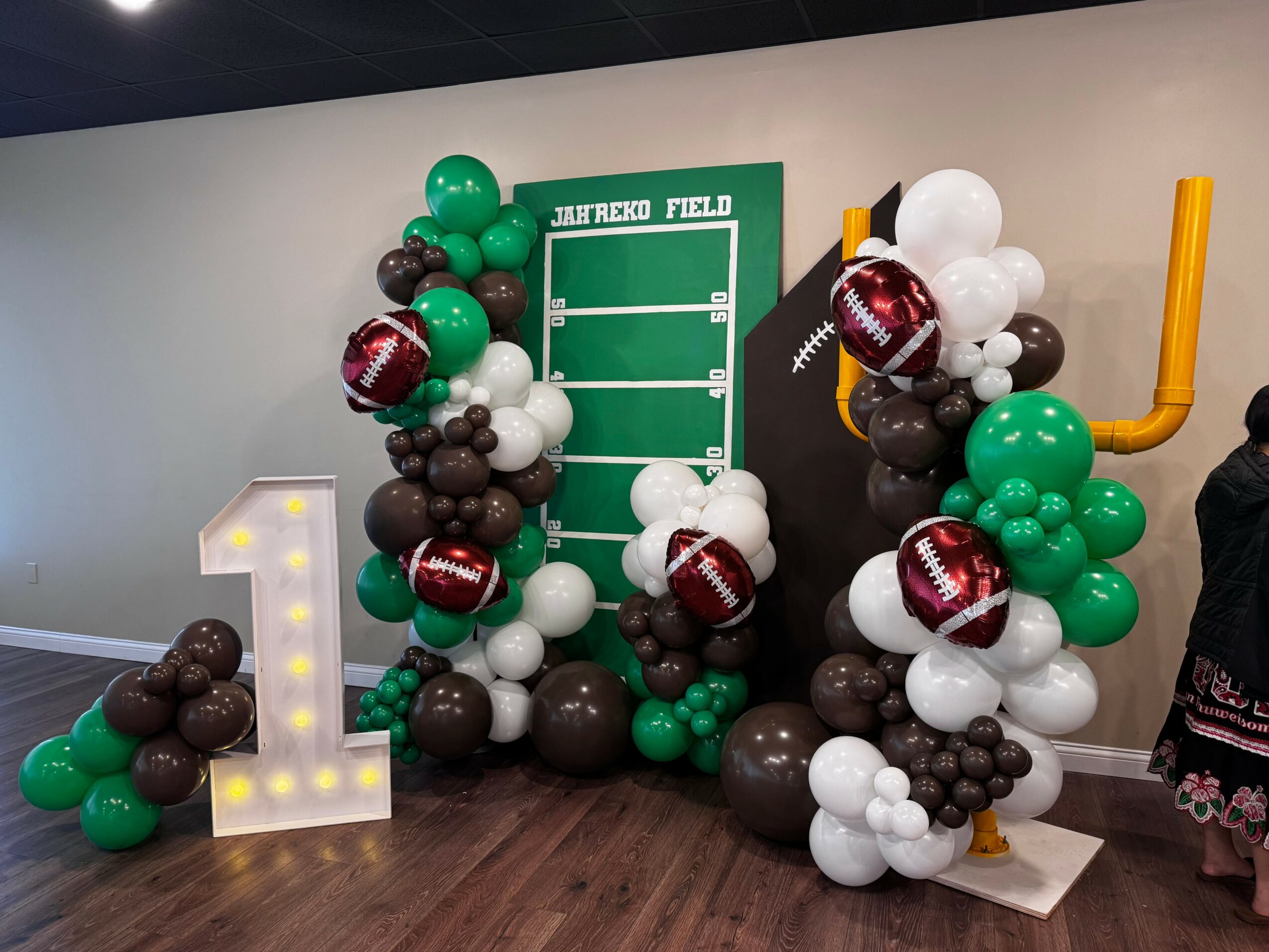 Decorative balloon arrangement with a large illuminated number one, a green scoreboard, and a yellow baseball bat, celebrating a baseball-themed event.