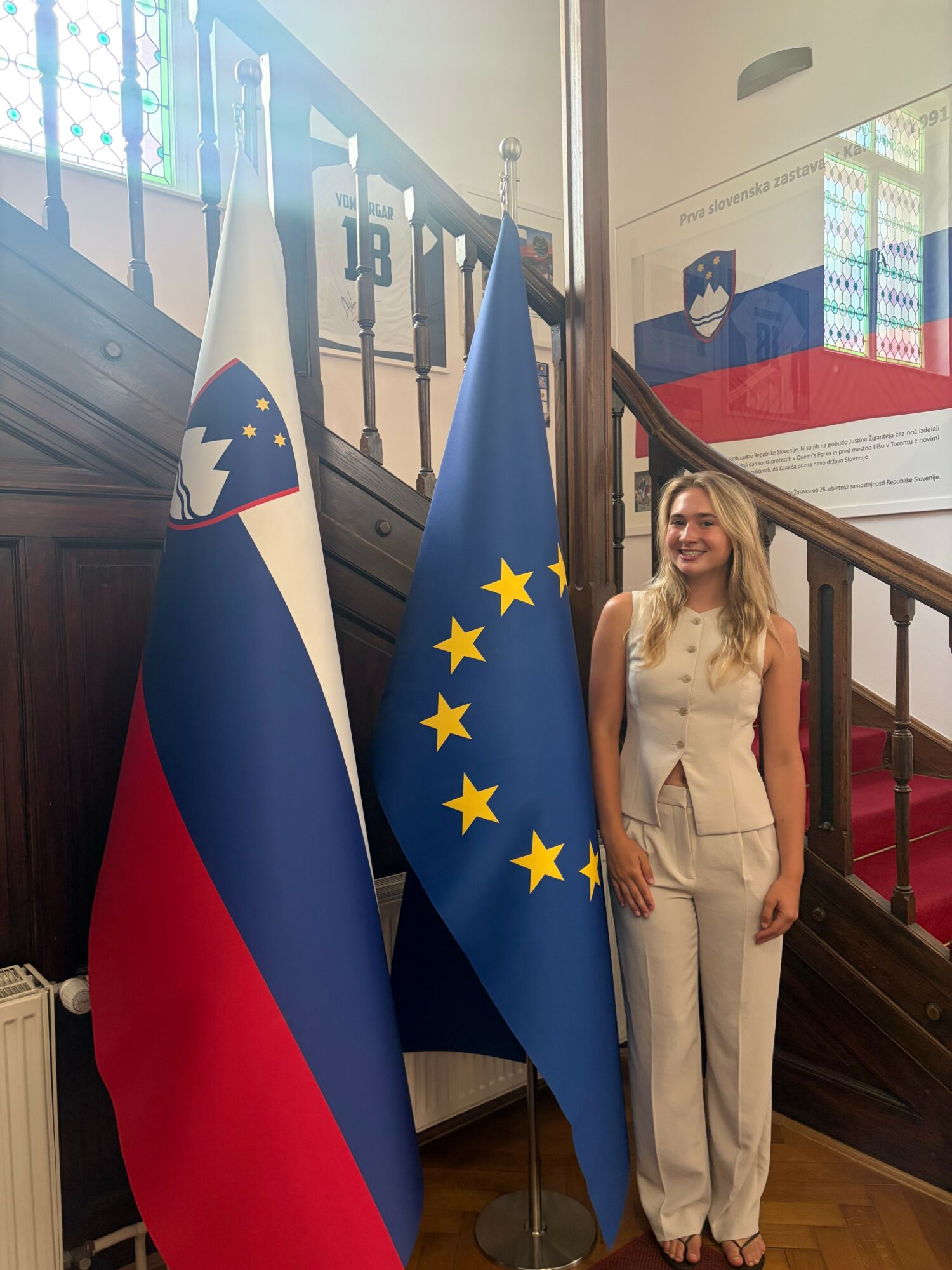 Woman in white dress standing next to European Union and Slovenian flags inside a building.