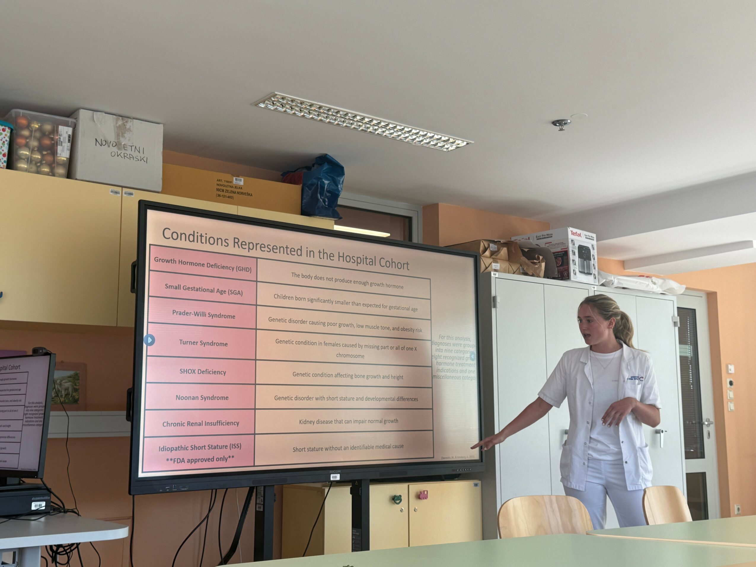 A woman in medical attire points at a large screen displaying hospital conditions, in a room with cabinets and storage boxes.