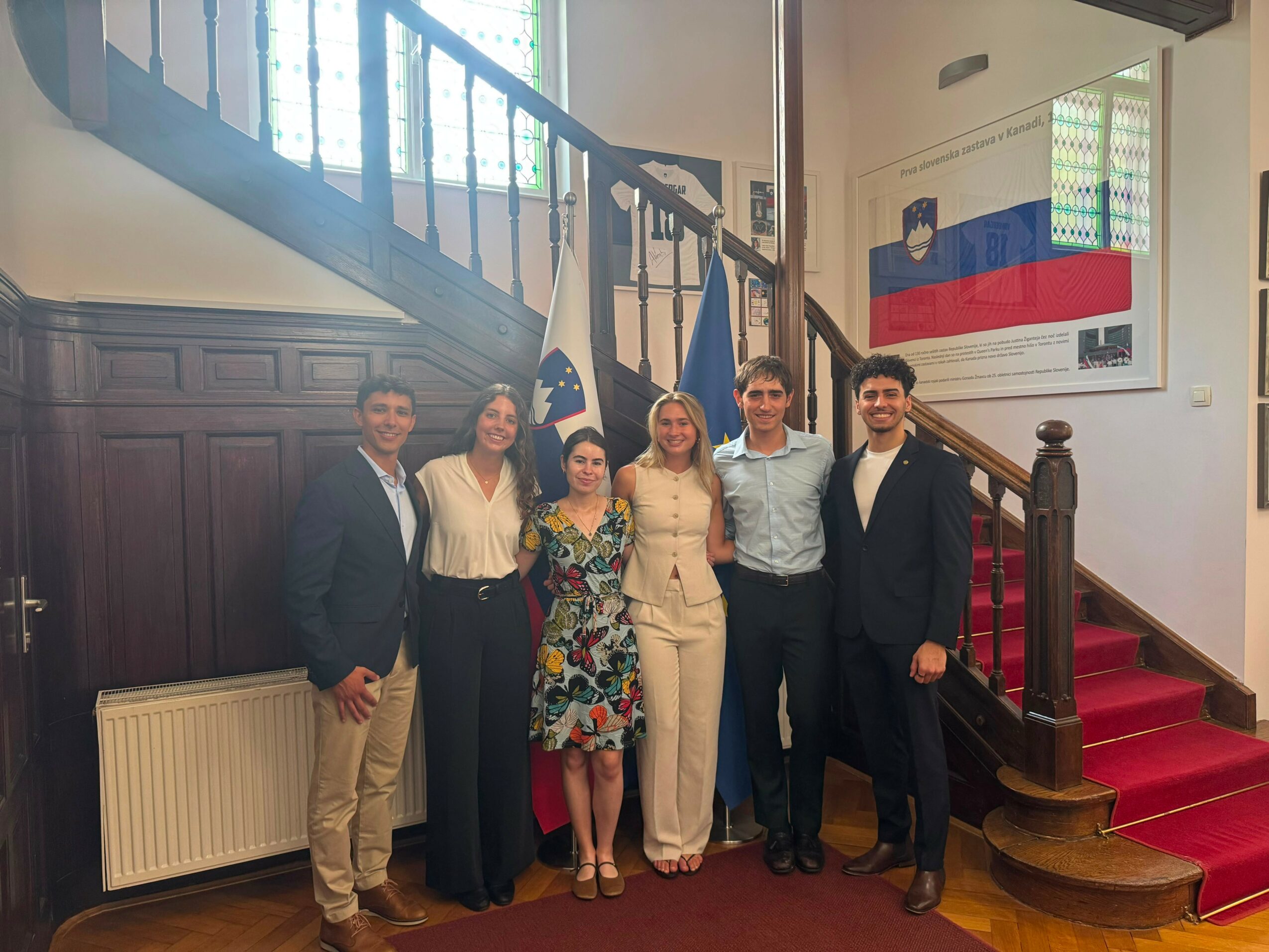 Group of six people standing in front of a staircase with flags and a flag on the wall, smiling for the photo.
