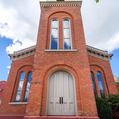 Front view of a red brick building with tall arched windows and a large central door, under a partly cloudy sky.