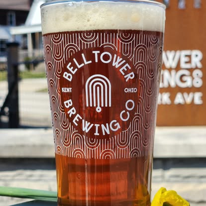 A pint glass filled with amber beer, featuring the Bell Tower Brewing Co logo, outdoors with a building and sign in background.