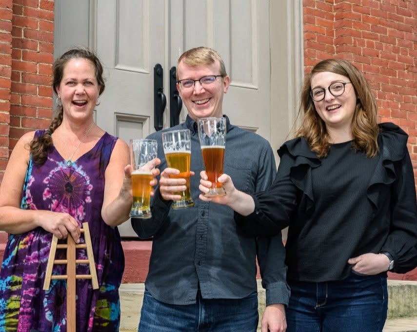 Three people smiling and holding glasses of beer outside a building with a brick wall and door.