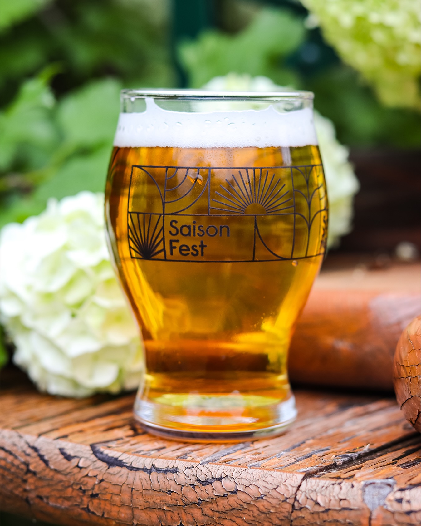 A glass of beer on a wooden surface with green and white flowers in the background.
