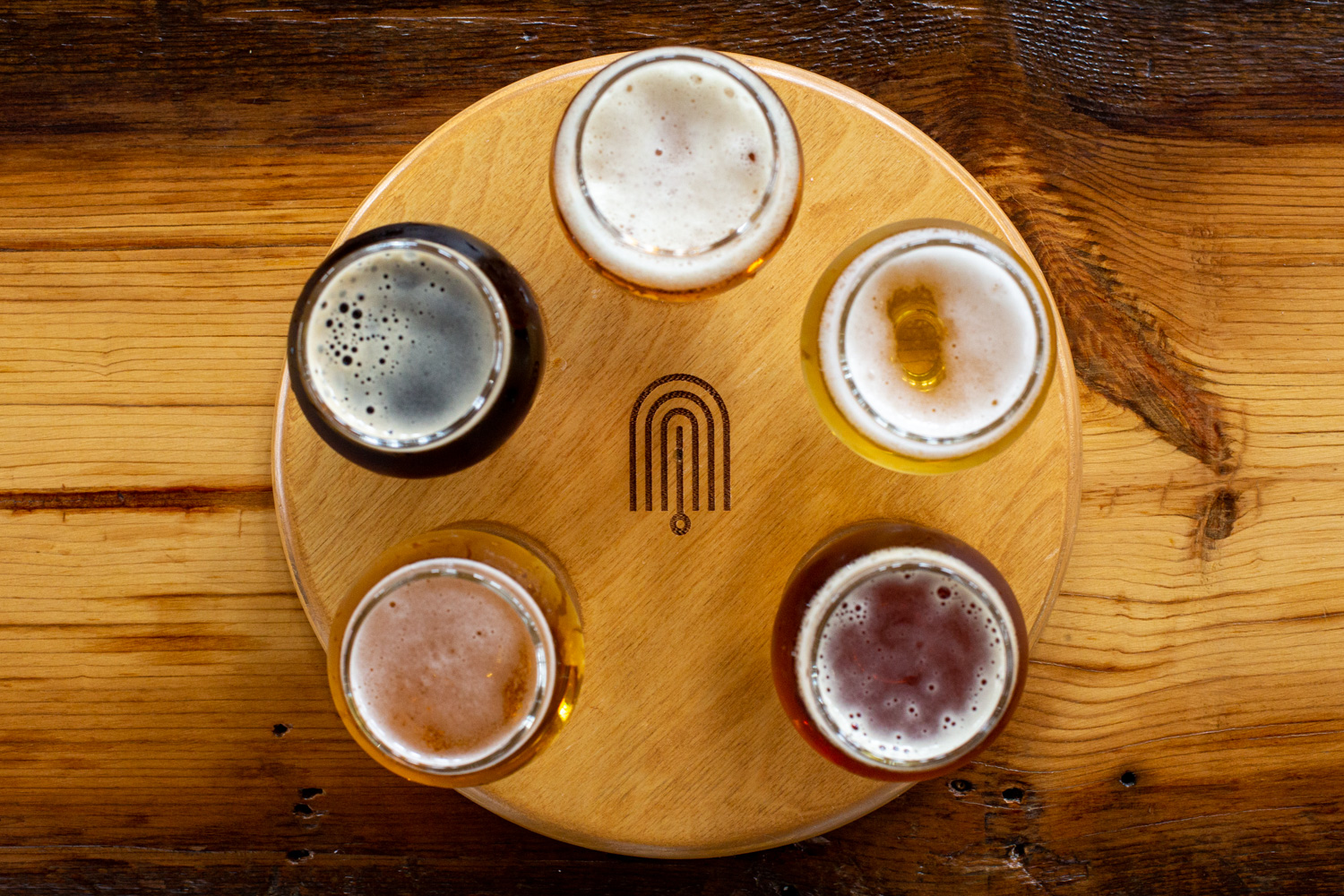 Six glasses of beer on a round wooden tray on a wooden table, viewed from above.