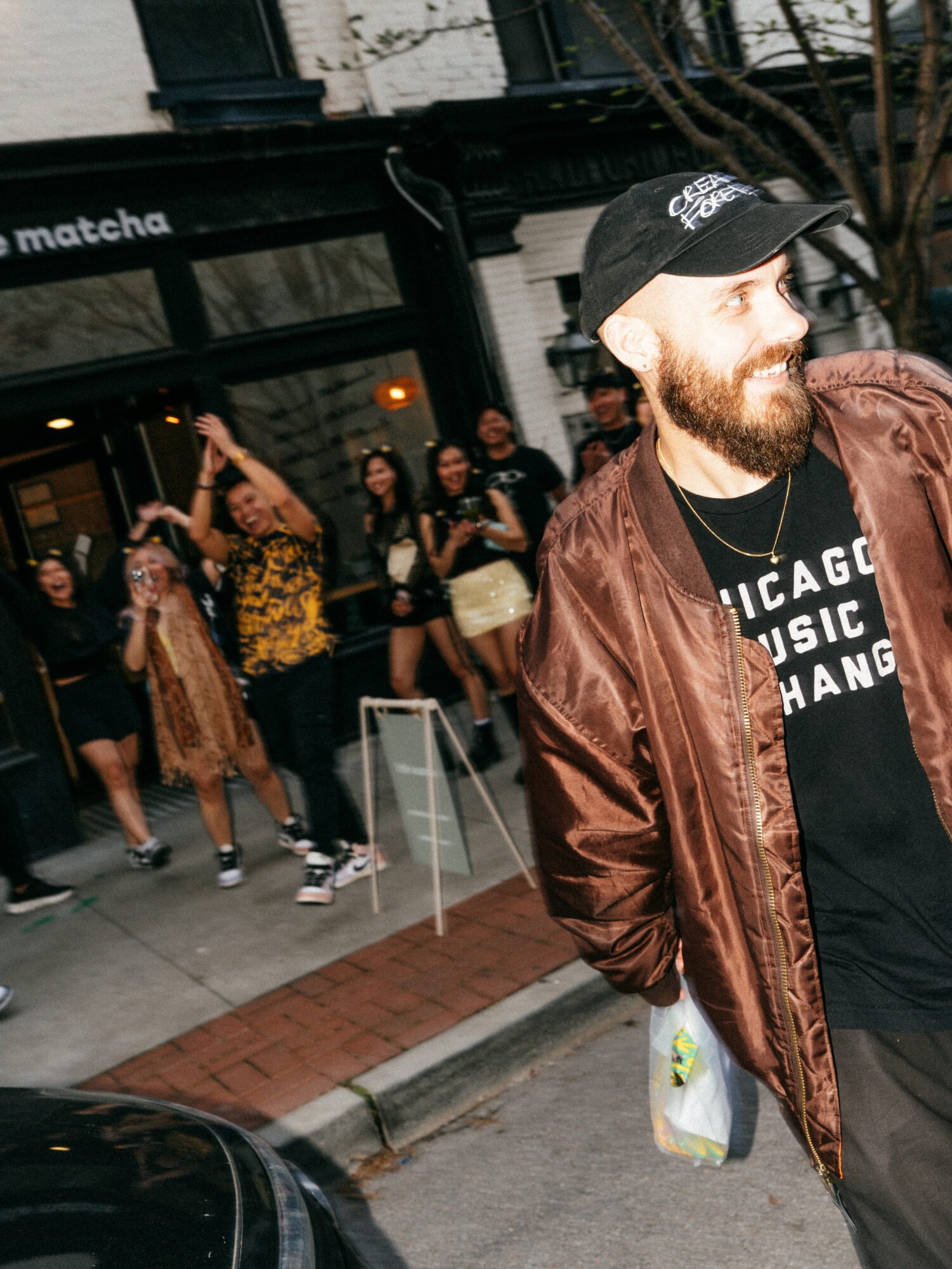 A bearded man wearing a black cap and brown jacket stands on a city street with a group of people in the background.
