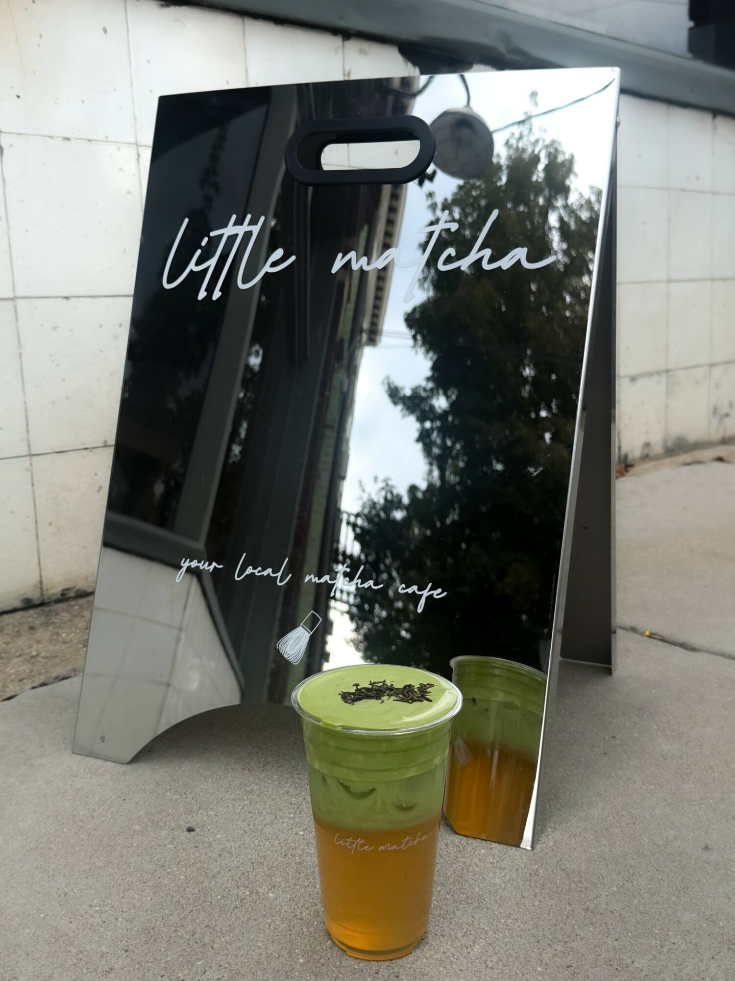 Mirror with handwritten text, reflection of trees, two green and orange drinks on ground.