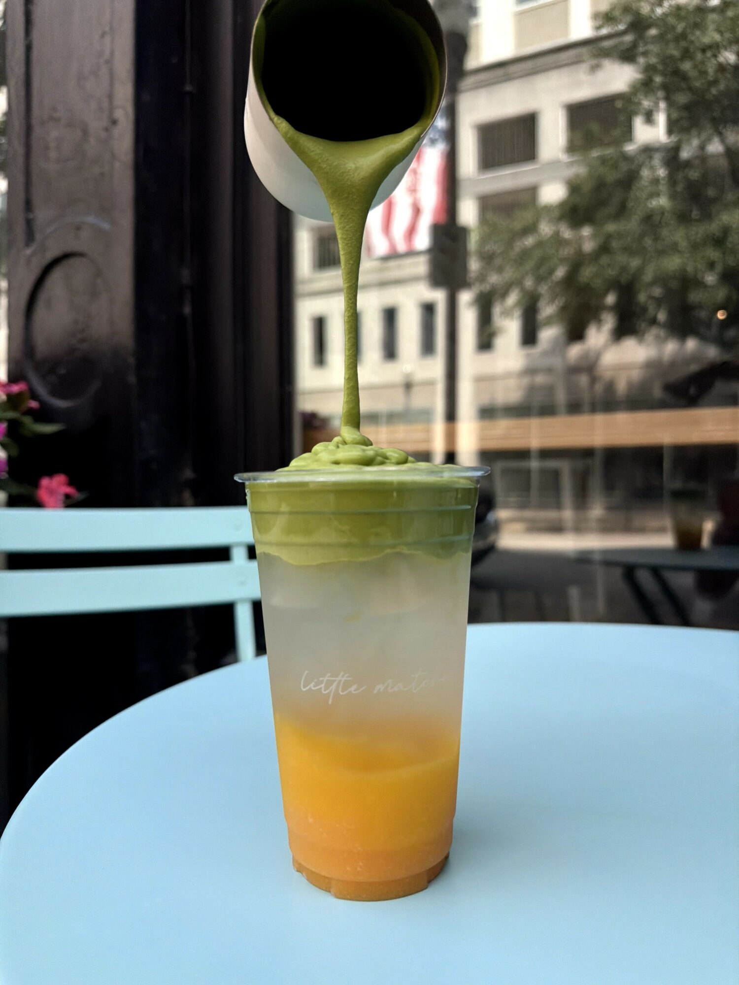 Green matcha latte being poured into a clear cup with layered yellow and orange at the bottom, on a blue table.