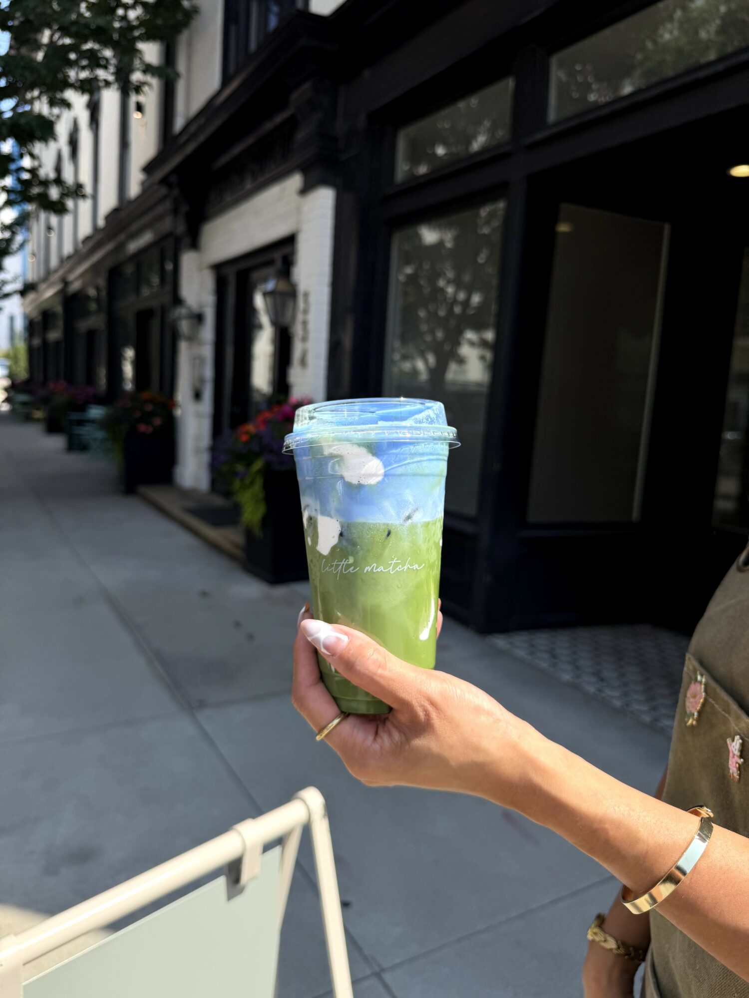 Person holding a colorful drink in a clear cup outdoors on a city sidewalk, with buildings and trees in the background.