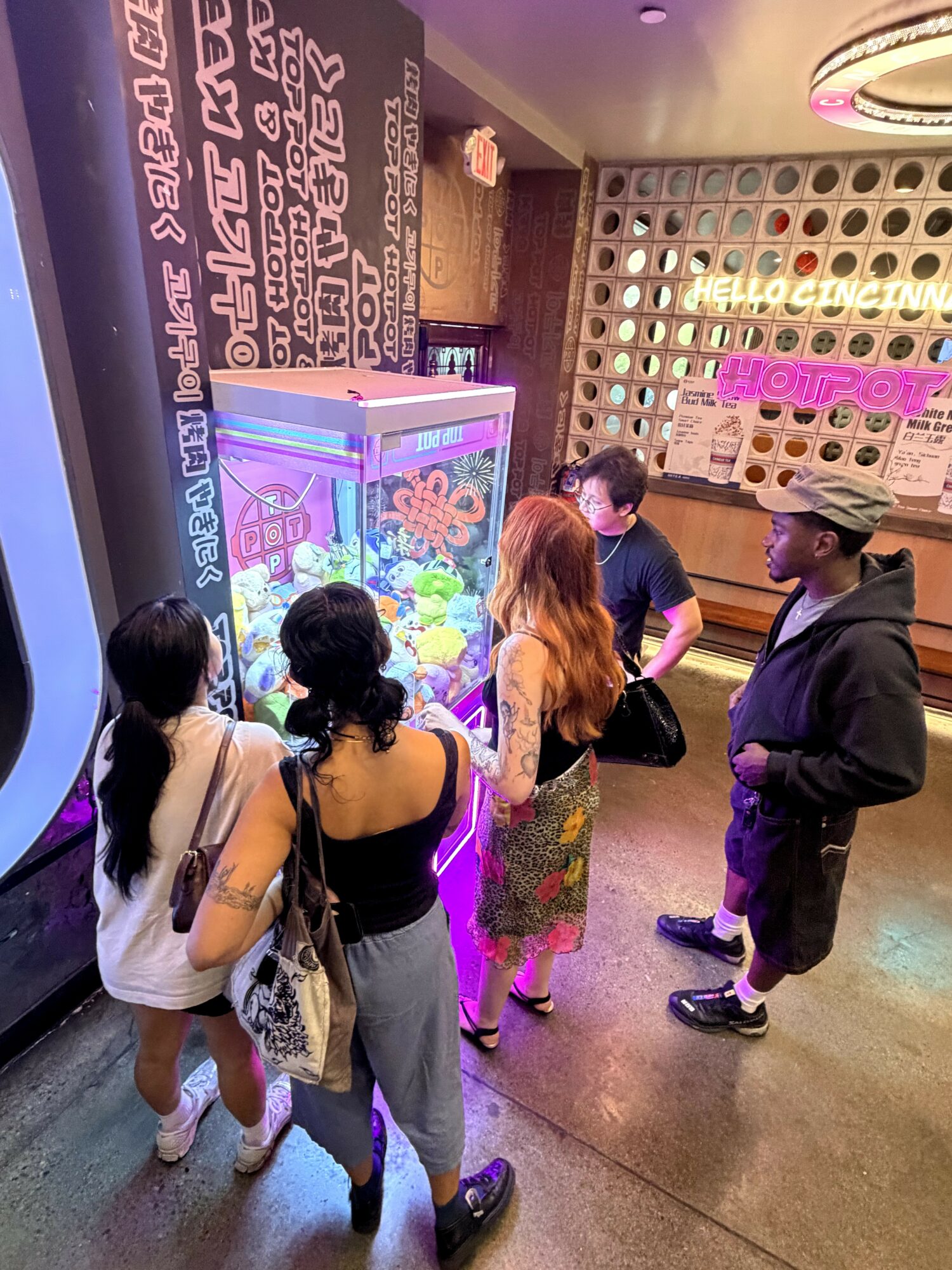Group of five people gathered around a claw machine in an arcade, with colorful lights and patterned wall behind.
