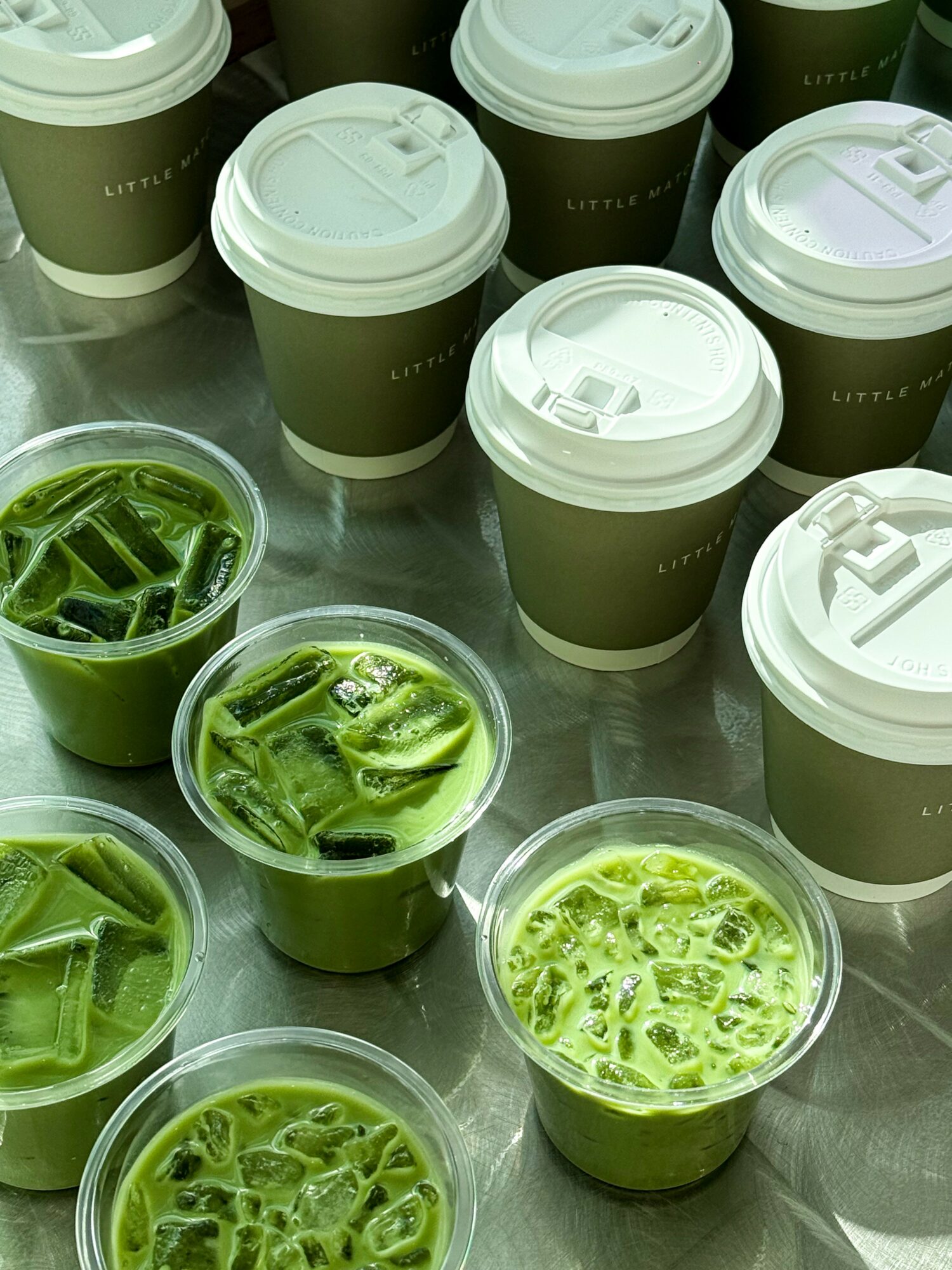 Multiple takeaway coffee cups with white lids and green drinks with ice, on a metallic surface.