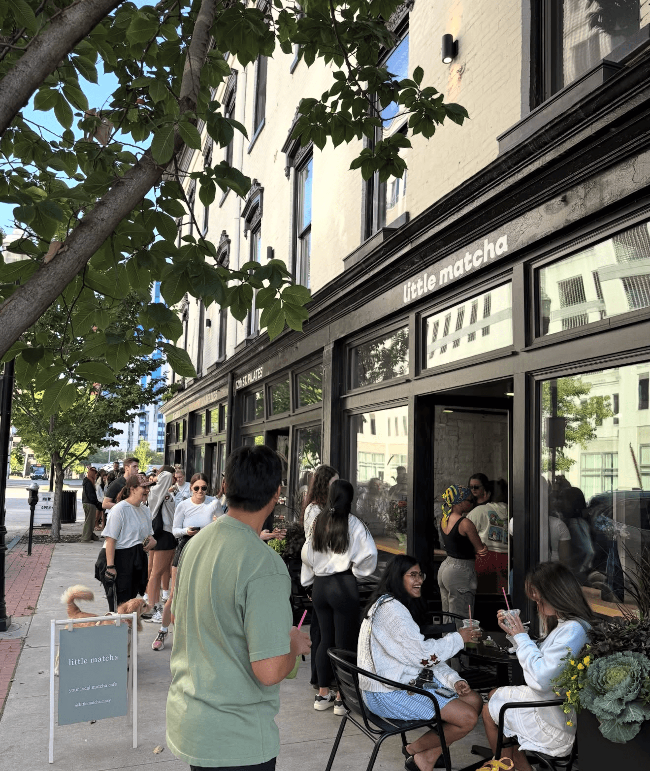 People gather outside a storefront with large windows, some sitting and others standing, with trees and buildings in the background.