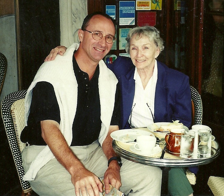A man and an elderly woman sitting together at a table, smiling, with cups and dishes on the table.