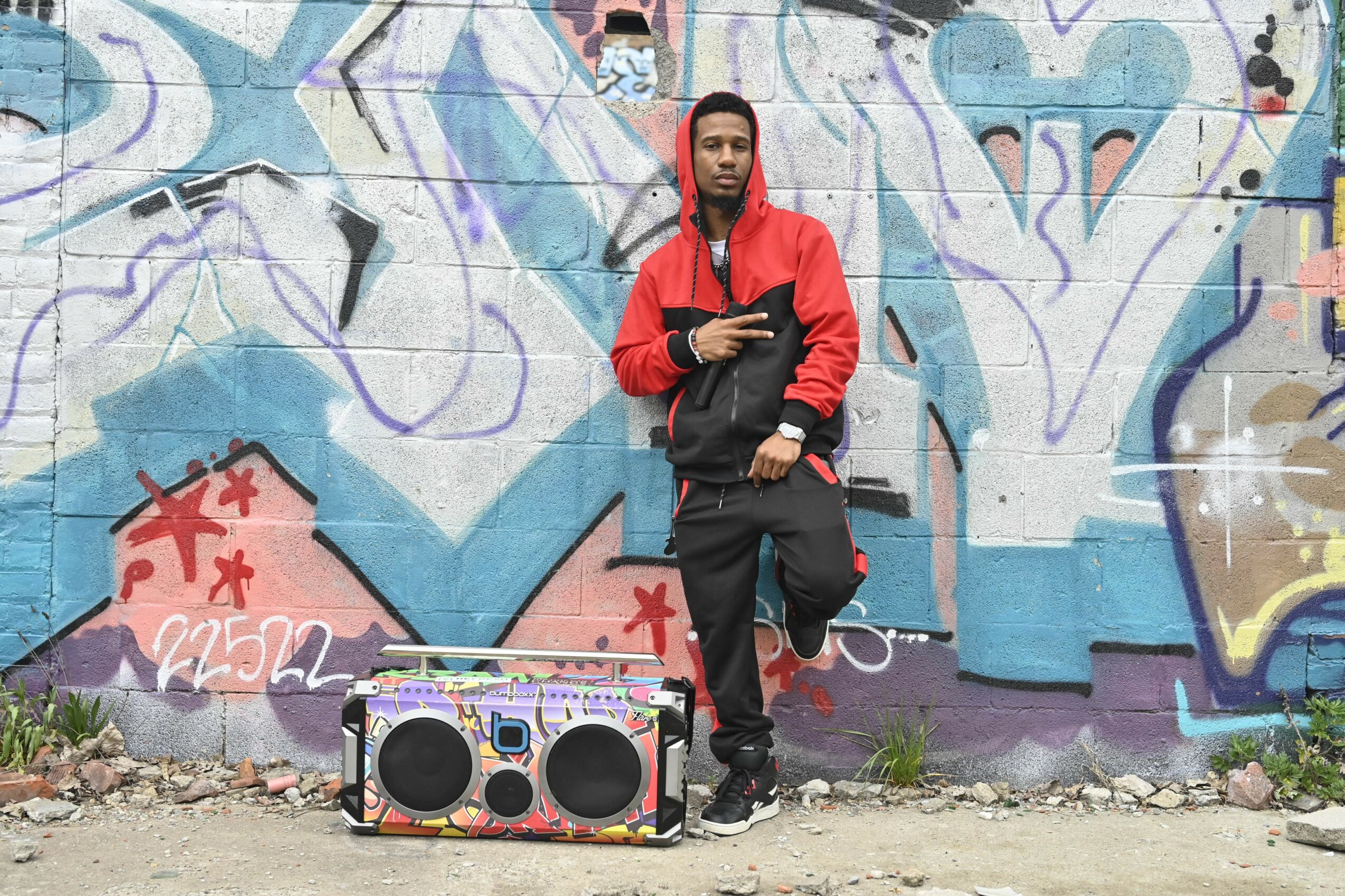 Person in red and black jacket standing in front of graffiti wall with boombox on ground, holding a phone.