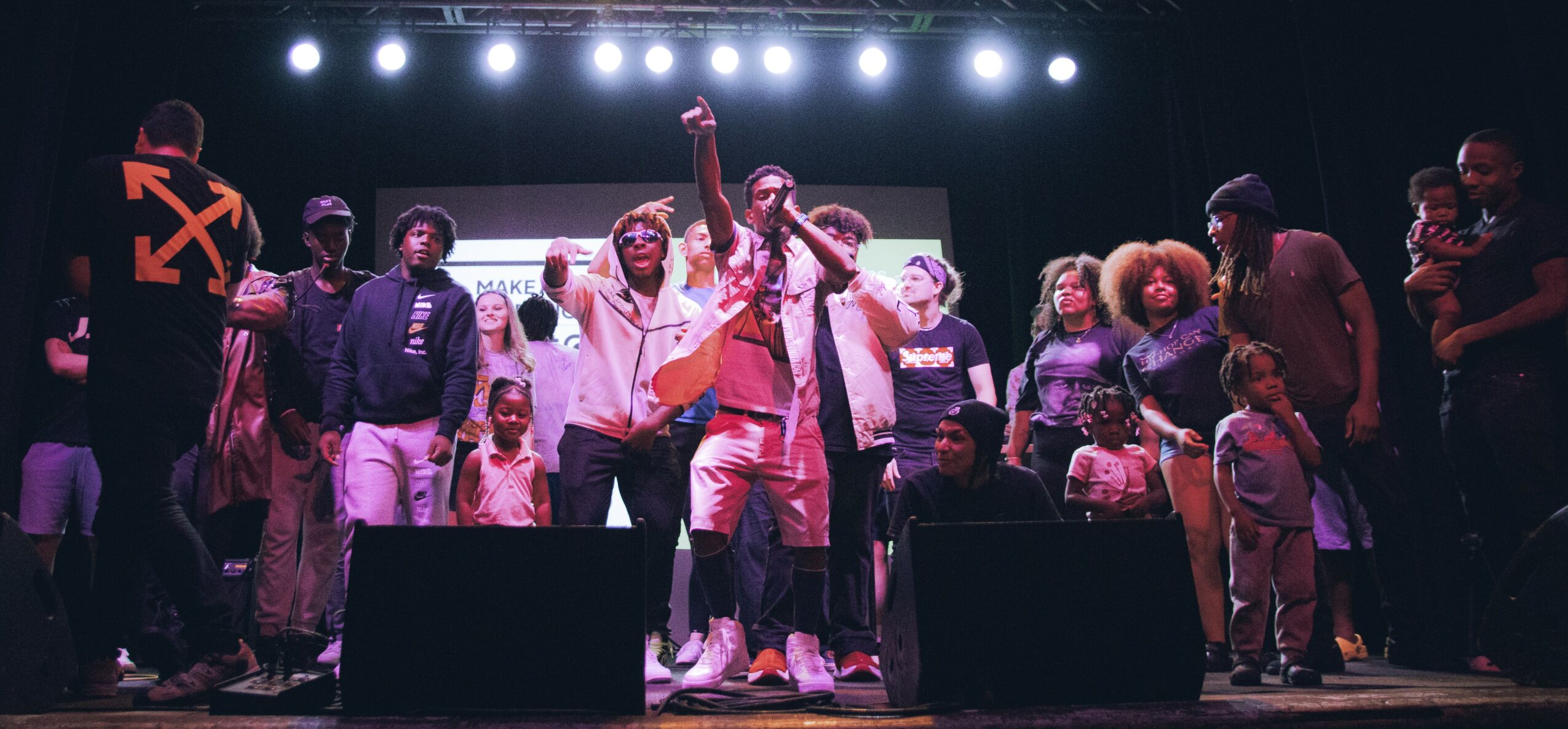 Group of people on stage, some raising hands, with stage lights overhead, diverse in age and appearance.
