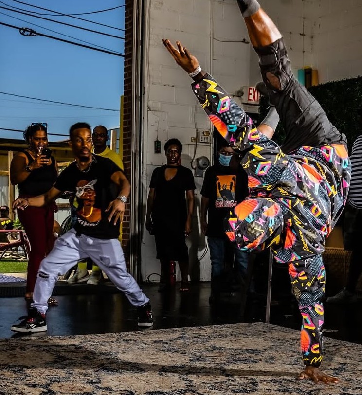 Person performing a handstand on one hand, wearing colorful clothing, with a group watching indoors. Word count: 20.