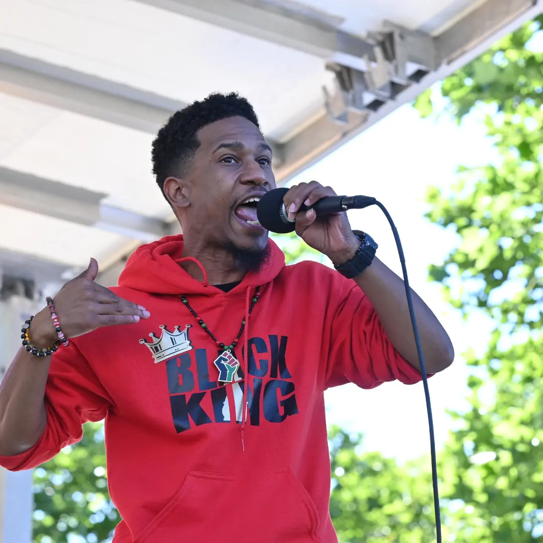 Person singing into a microphone outdoors, wearing a red hoodie with text, with trees and sky in background.