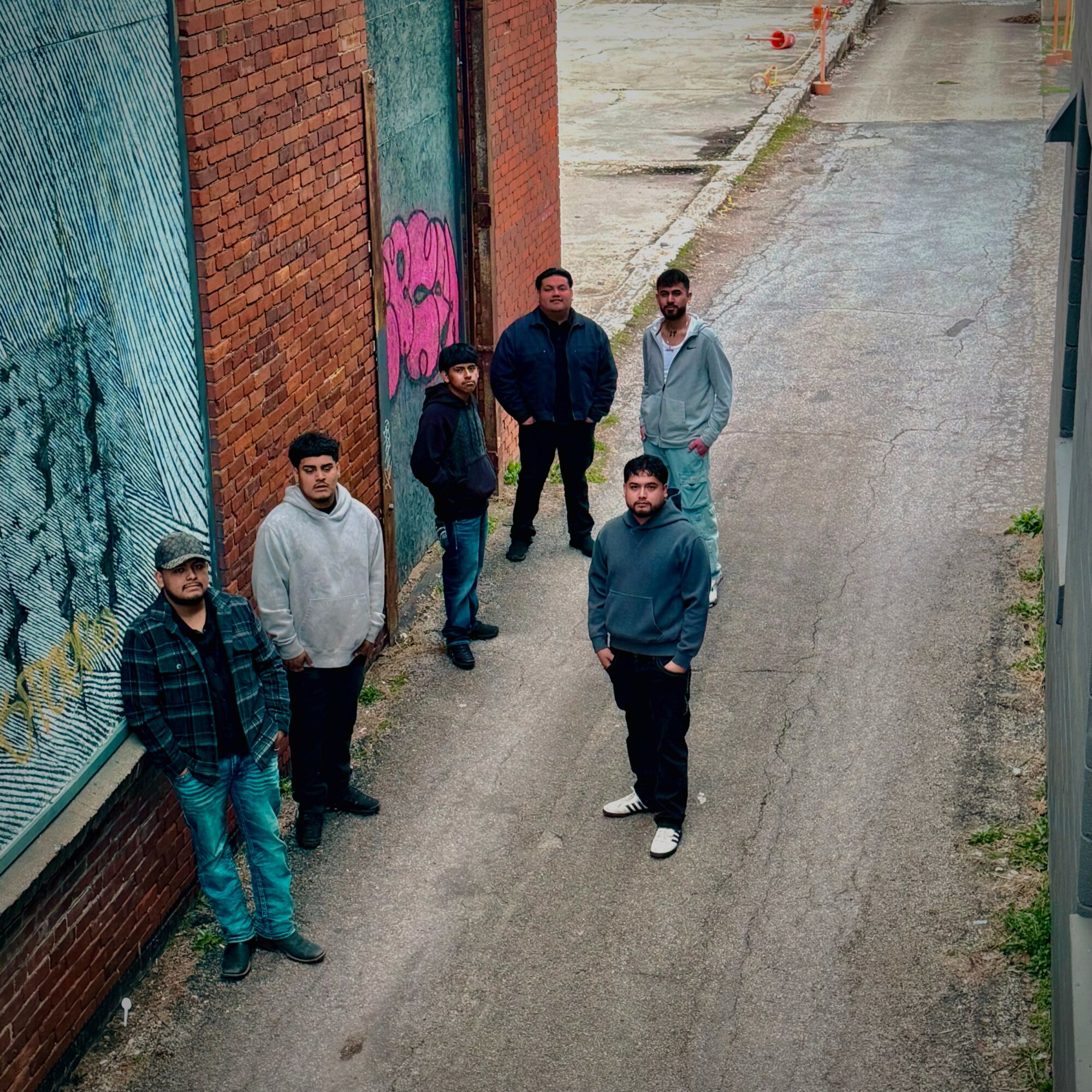 Six people standing on a narrow street next to a brick wall with graffiti, viewed from above.
