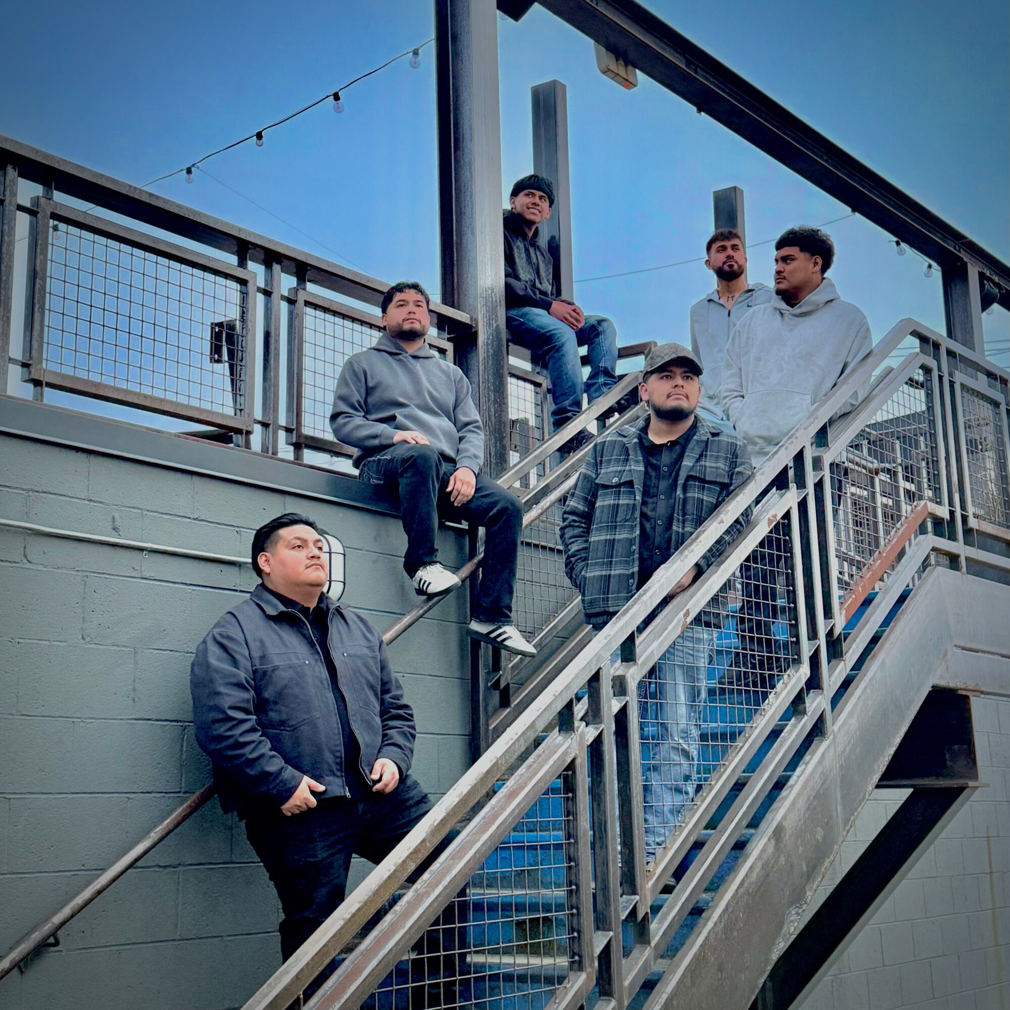 Six men standing on an outdoor staircase and platform, some leaning on the railing, against a blue sky background.