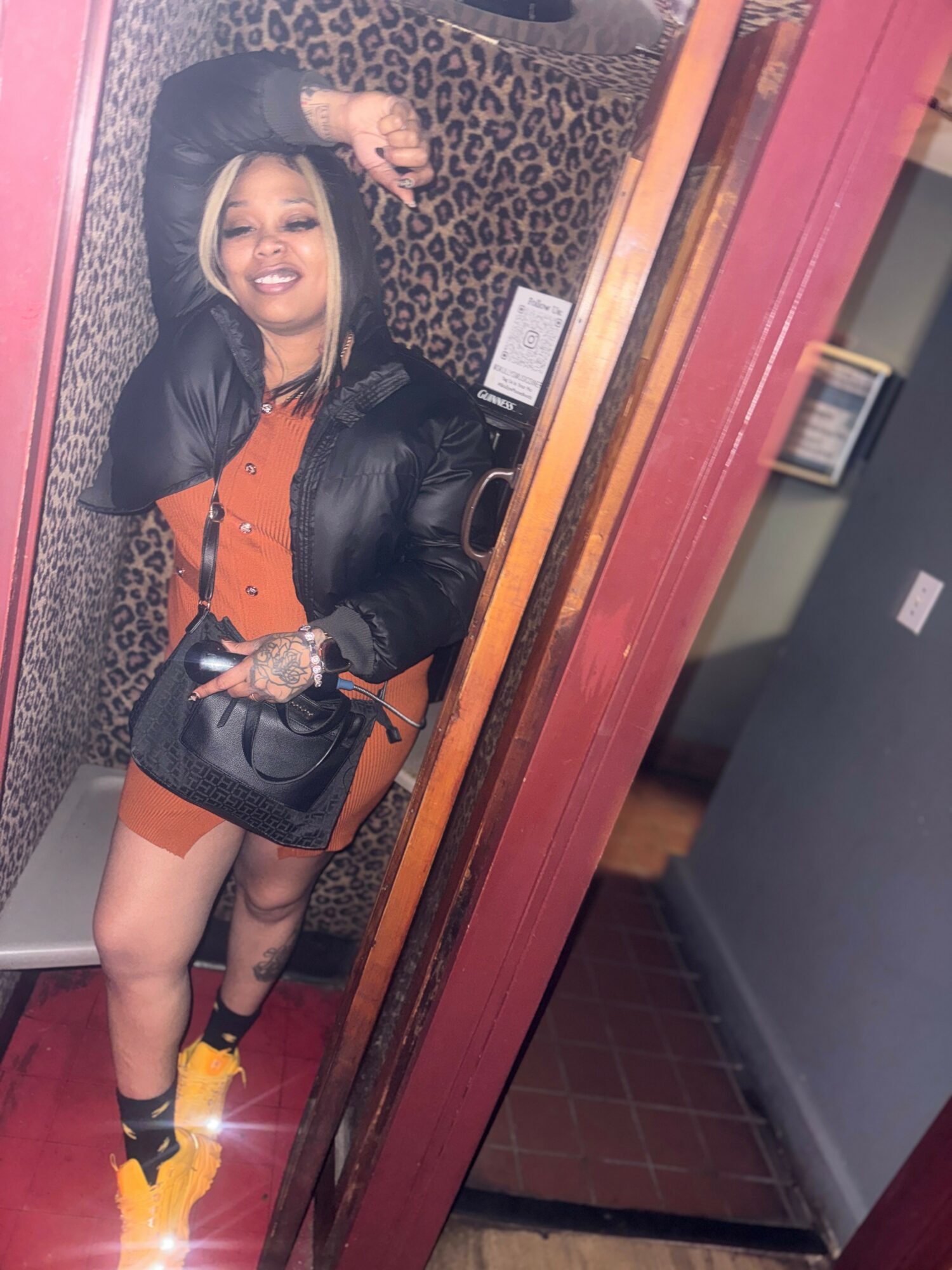 Person smiling, wearing a black jacket, orange dress, yellow shoes, taking a mirror selfie in a room with leopard print wall.
