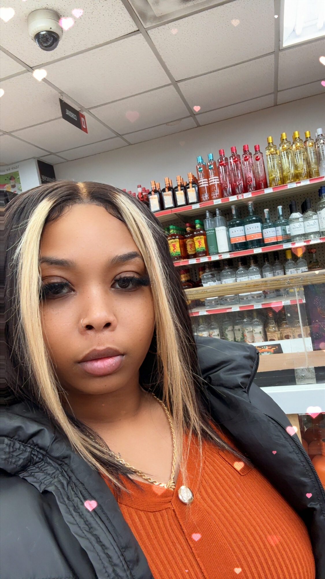 Young woman with blonde highlights and dark hair, wearing an orange top and black jacket, standing in a store aisle.