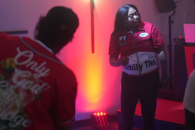Two women in red uniforms stand in a dimly lit room with colorful lighting, one facing forward and the other with back to camera.
