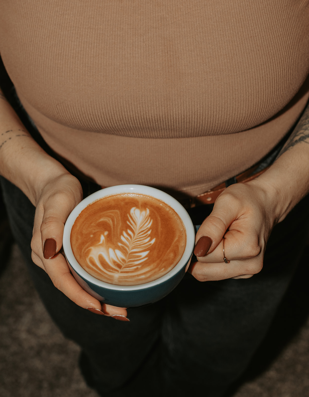 Person holding a cup of coffee with latte art, wearing a beige top and dark pants.