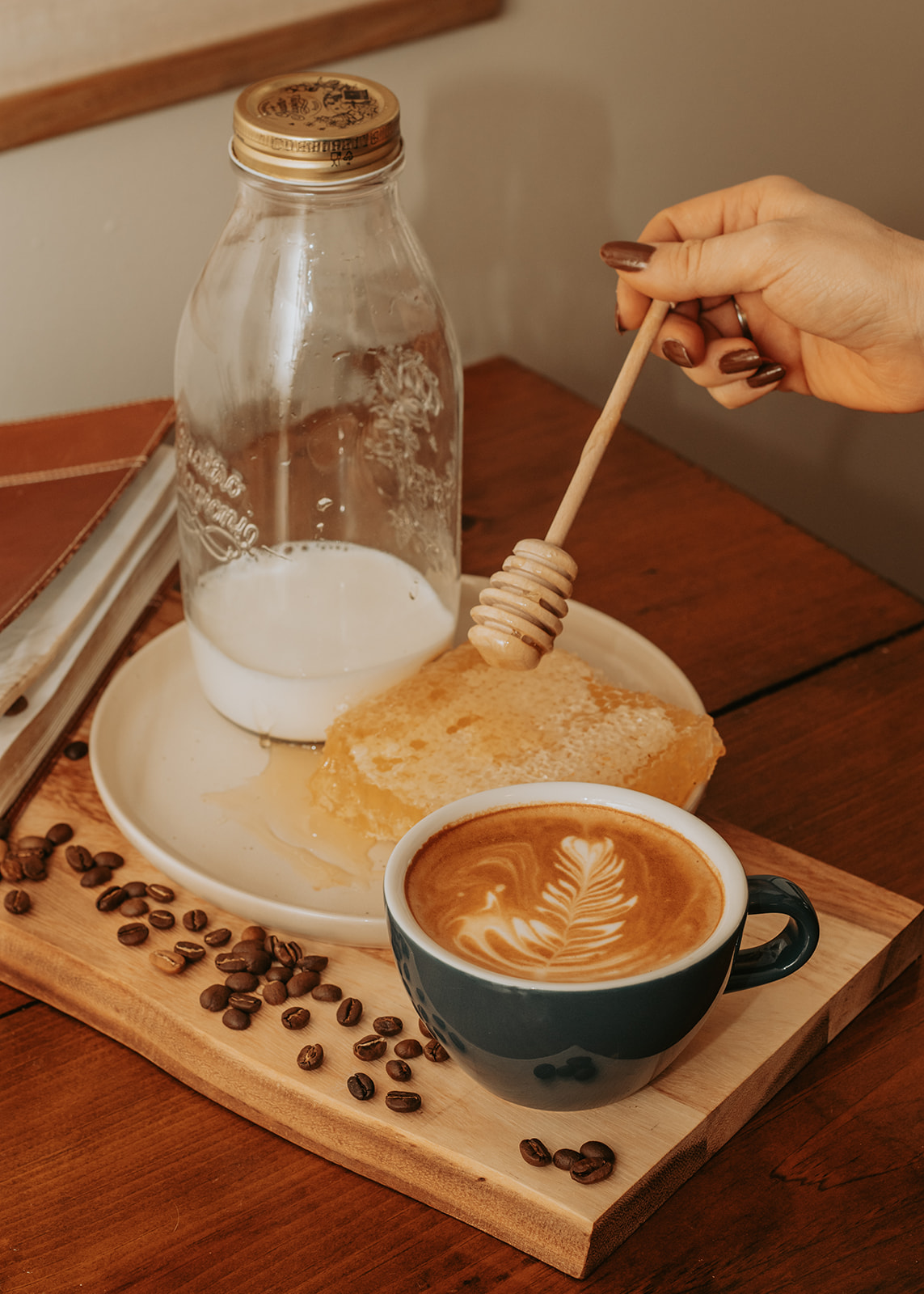 Hand holding a honey dipper over a plate with bread, coffee cup, coffee beans, and a glass bottle of milk on a wooden tray.