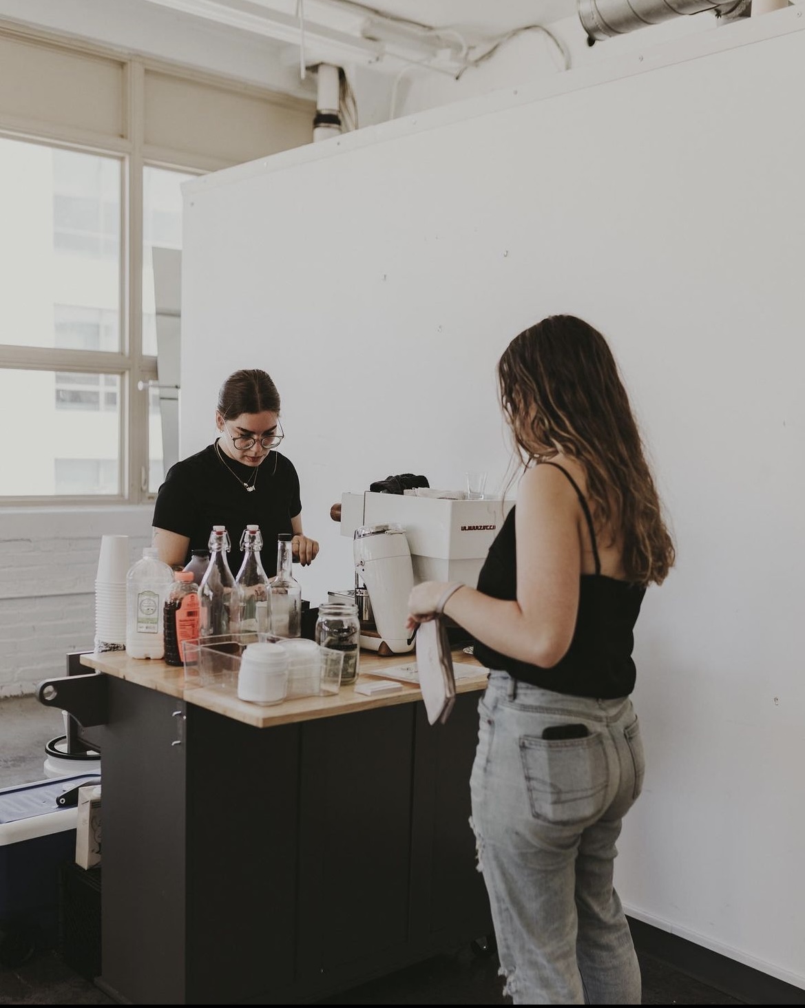 Two women in a room with large windows, one preparing drinks and the other looking at a phone, with a white wall background.