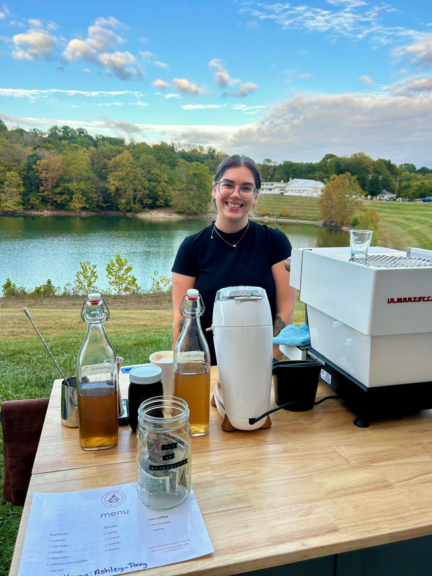 Smiling woman sitting at a table outdoors near a lake with trees and sky in background.