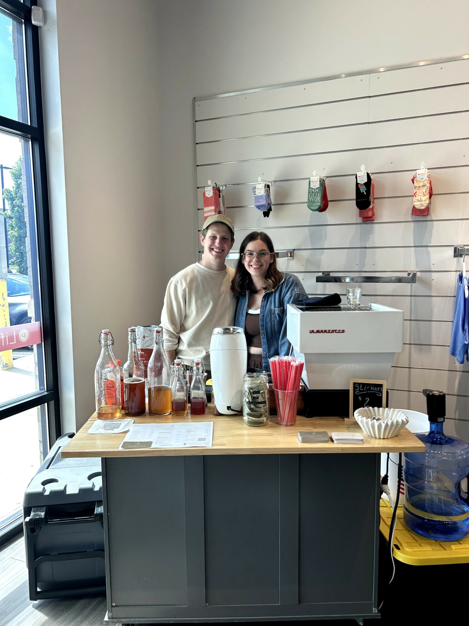 Two women stand behind a coffee counter with syrup bottles, a white coffee machine, and cups, in a cafe setting.