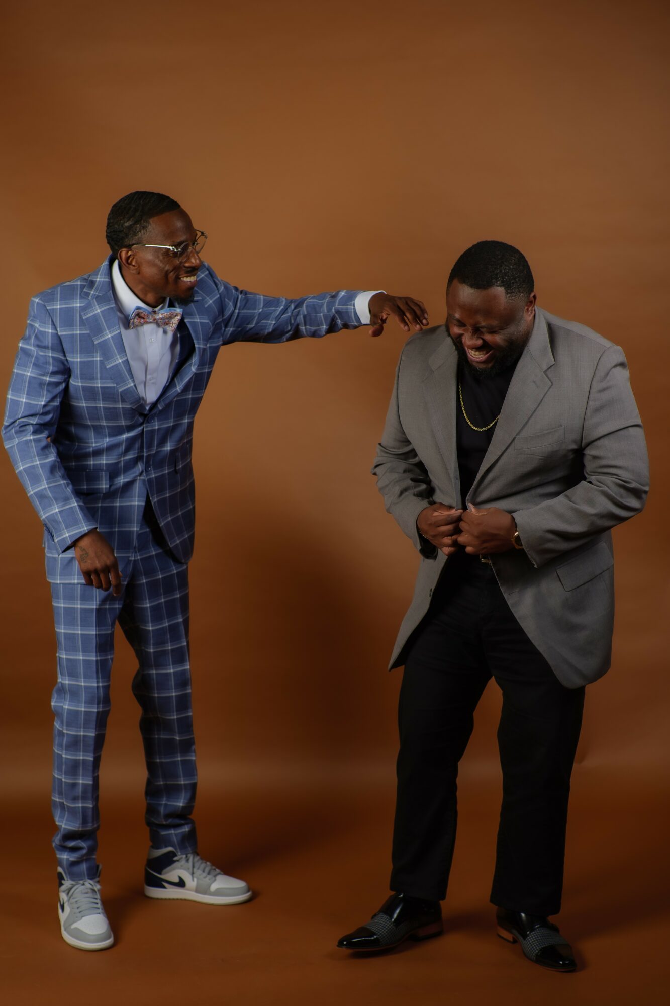 Two men laughing and interacting, one in a blue checkered suit and the other in a gray blazer, against a plain brown background.