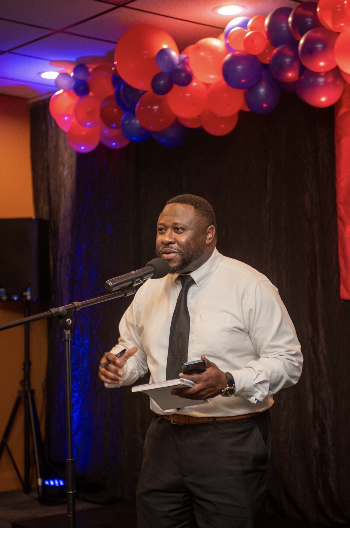 Man speaking into microphone on stage with red and purple balloons overhead, holding a tablet, in a formal setting.