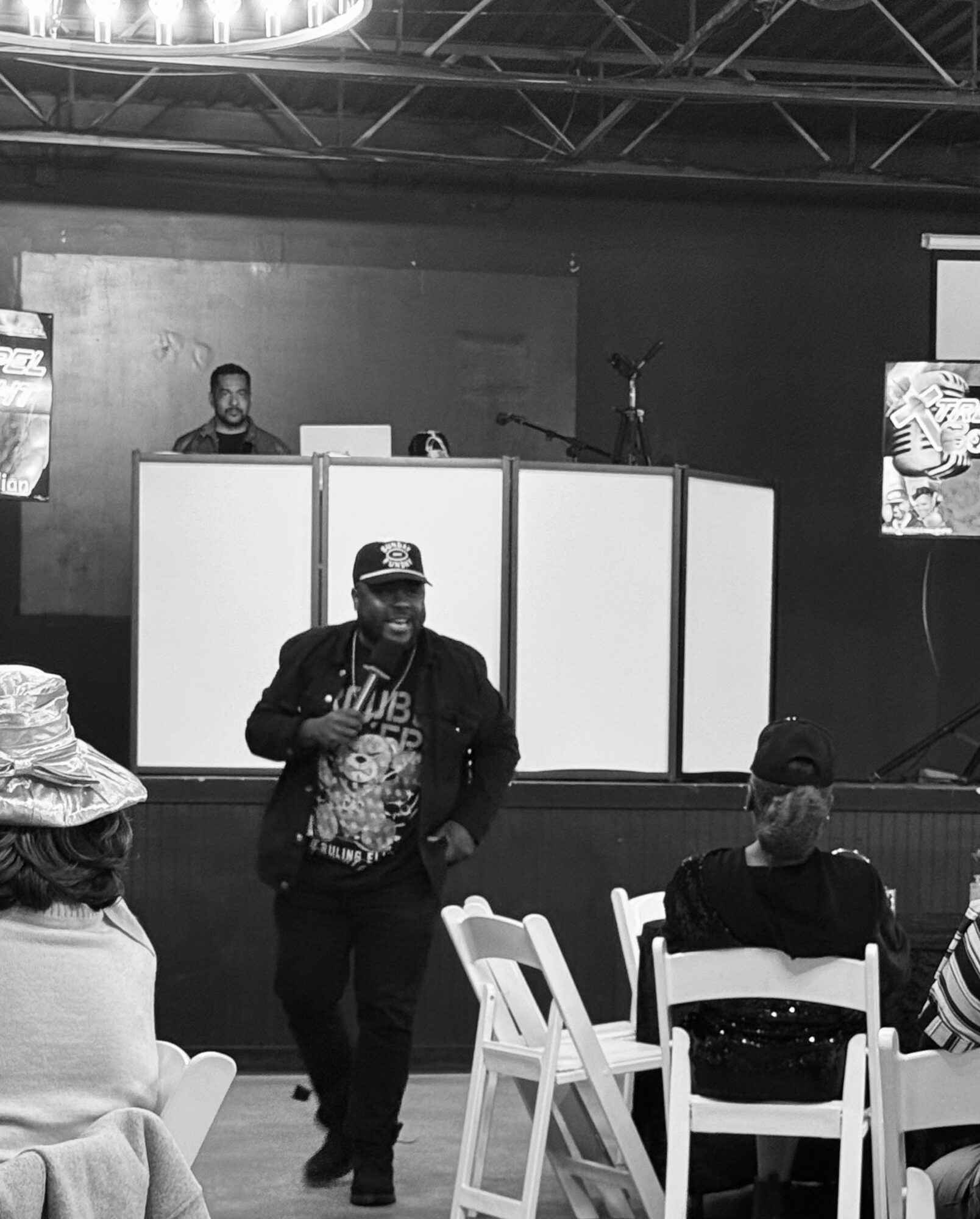 Person walking in front of seated audience at event with DJ booth and screens in background.