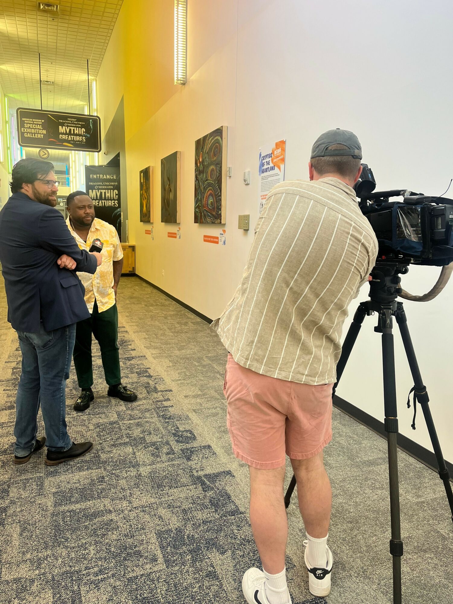 Two people standing in a hallway, one with a camera on a tripod, the other with a child, walls decorated with framed pictures.