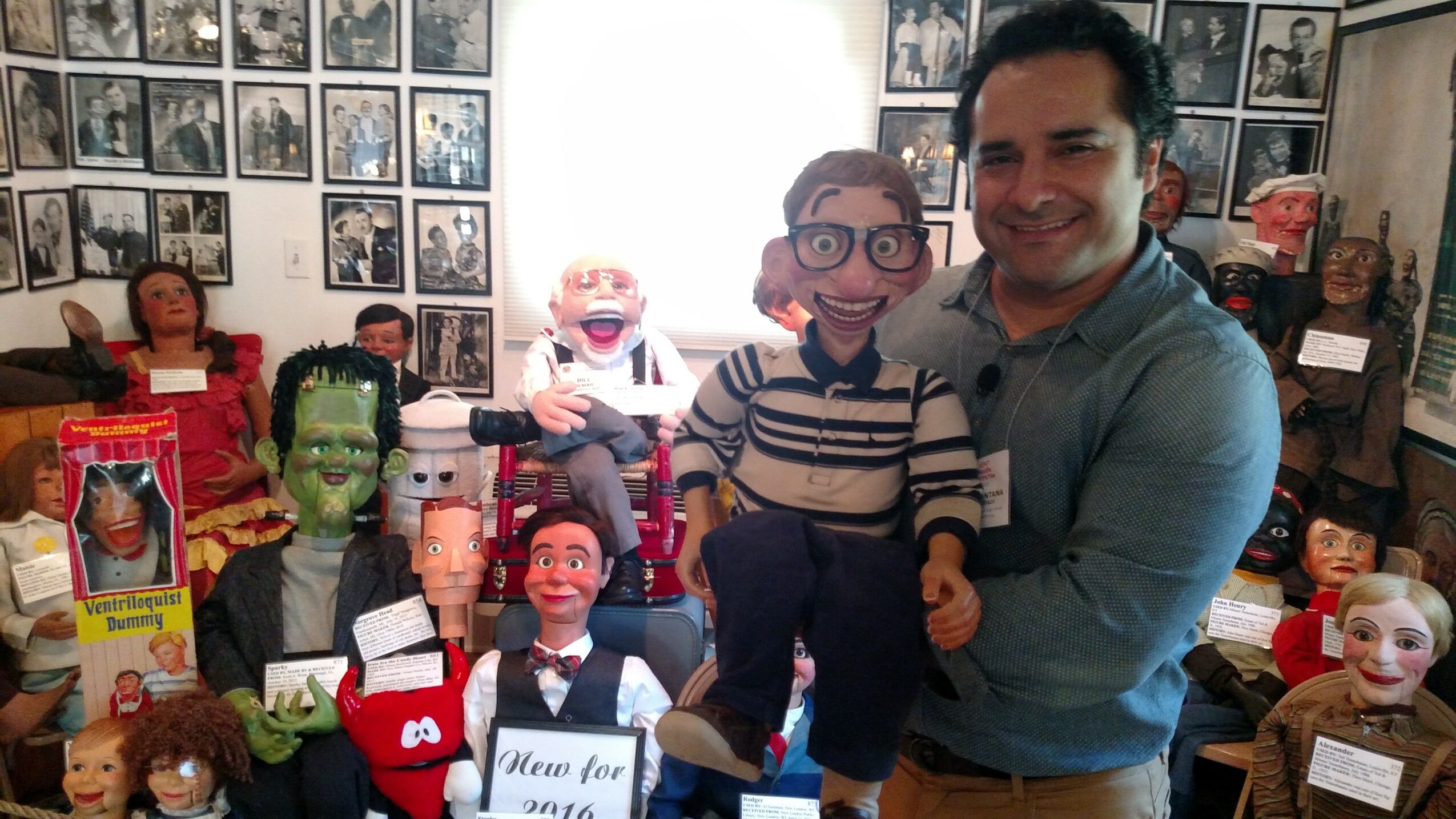 Man holding a puppet with glasses and striped shirt, surrounded by various puppets and photos on wall, in a room.