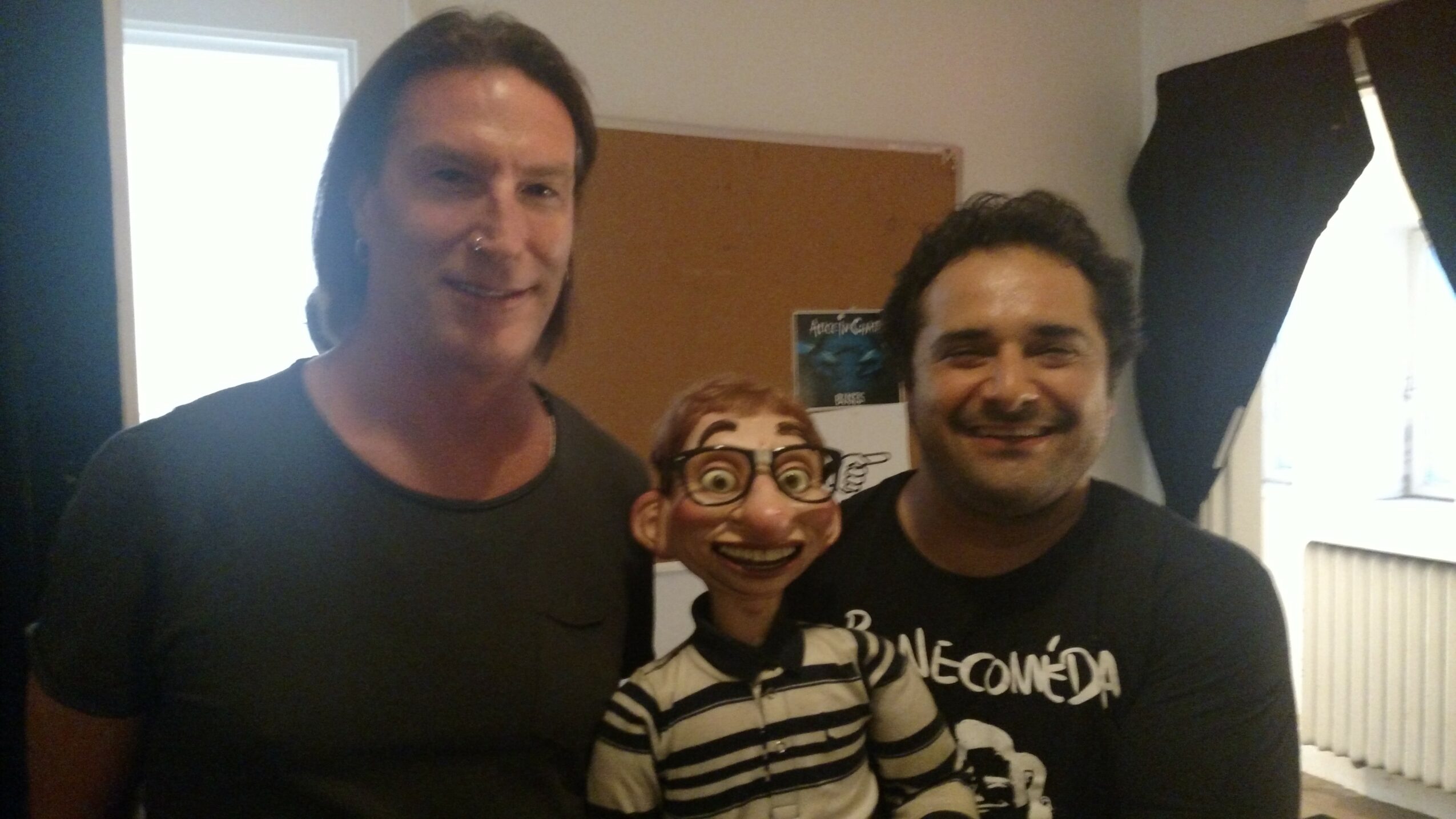 Two men standing indoors with a puppet between them, smiling, in a room with a window and curtains behind.