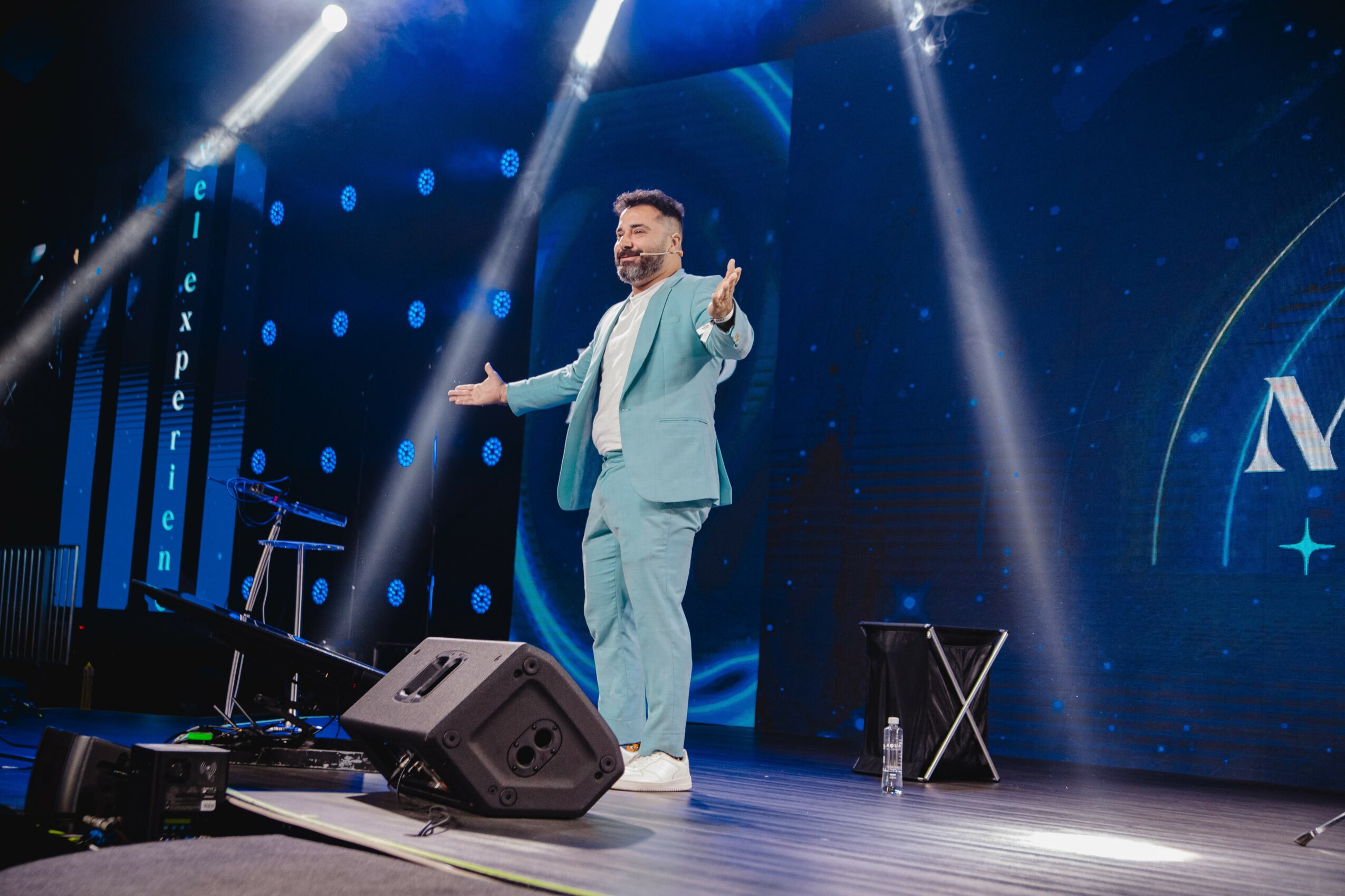 A man in a light-colored suit stands on stage with arms outstretched, under bright stage lights, with a large screen behind him.