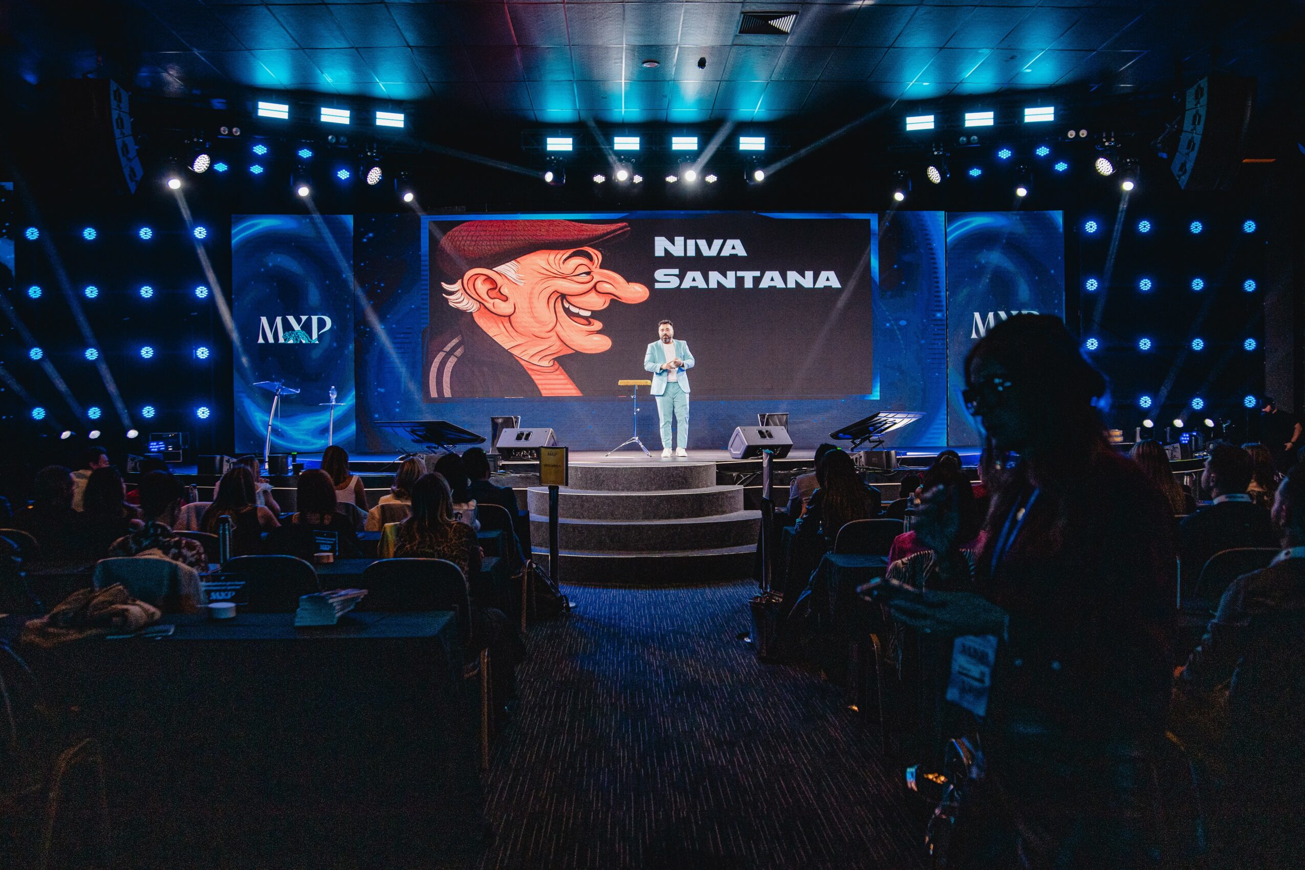 Person speaking on stage at a conference with a large screen displaying 'Niva Santana' and a caricature, audience seated.