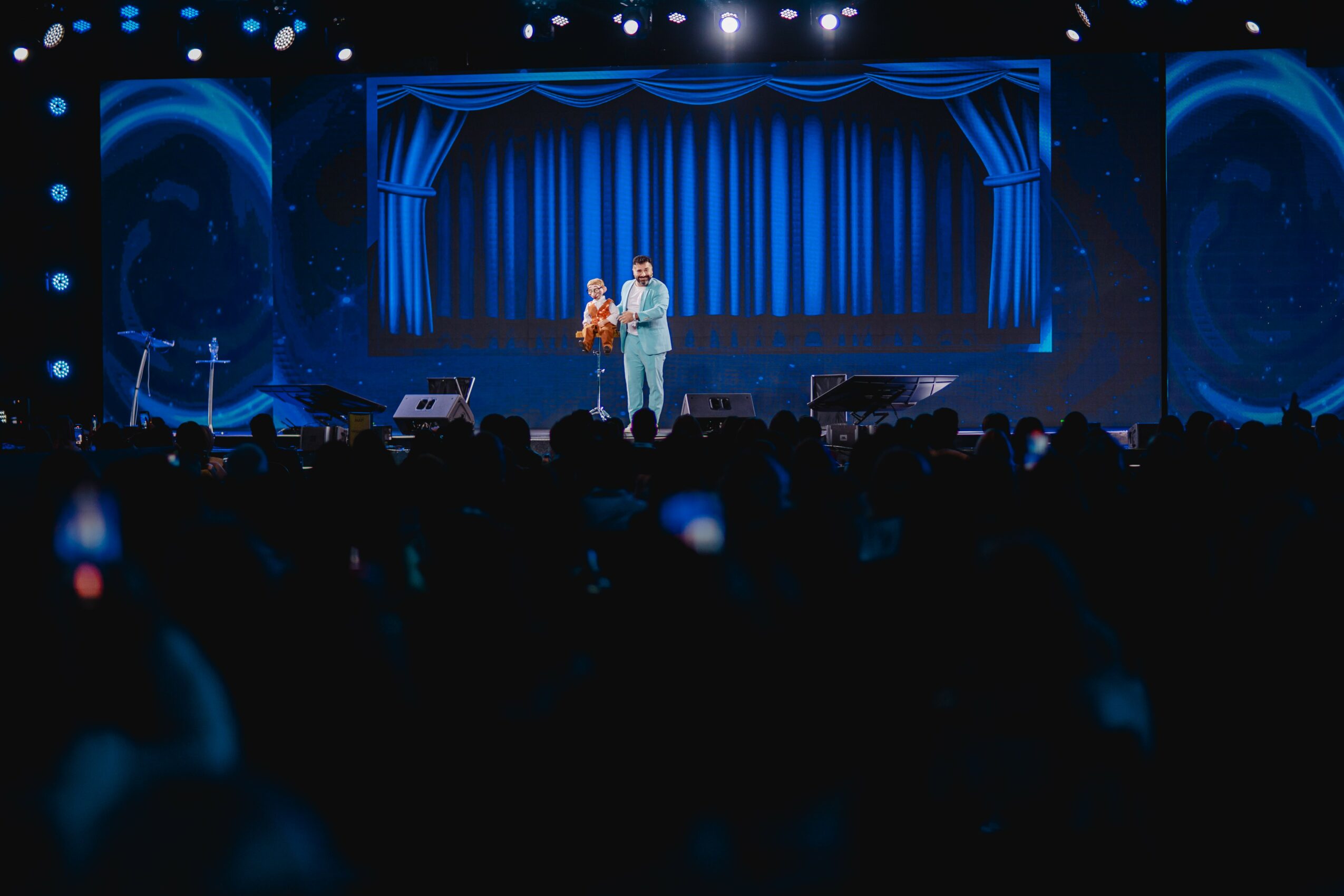 Two people stand on a stage with blue curtains and lighting, performing or speaking to an audience.