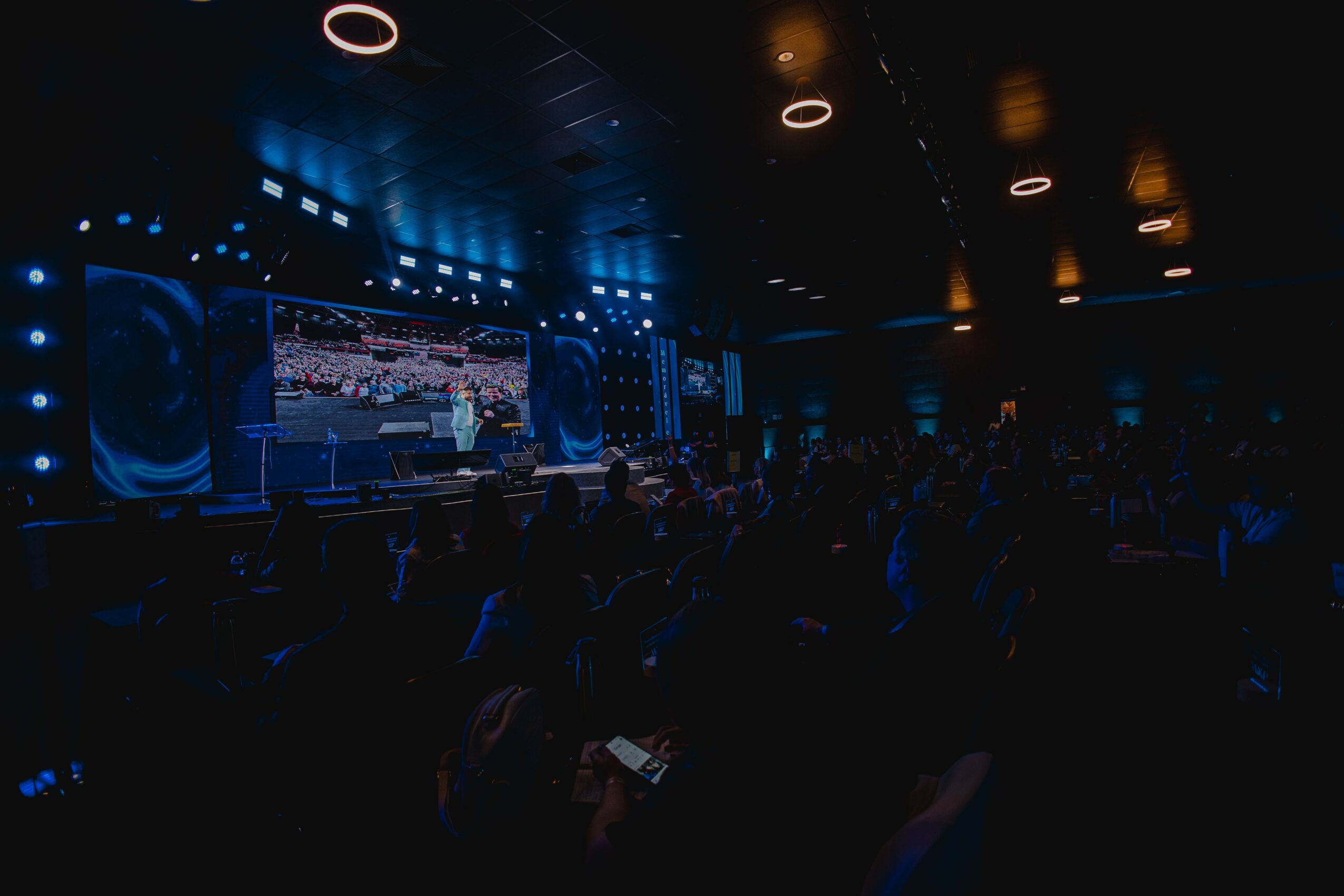 Stage with large screen and speakers in a dark auditorium, audience seated facing the stage, dim lighting, blue accents.