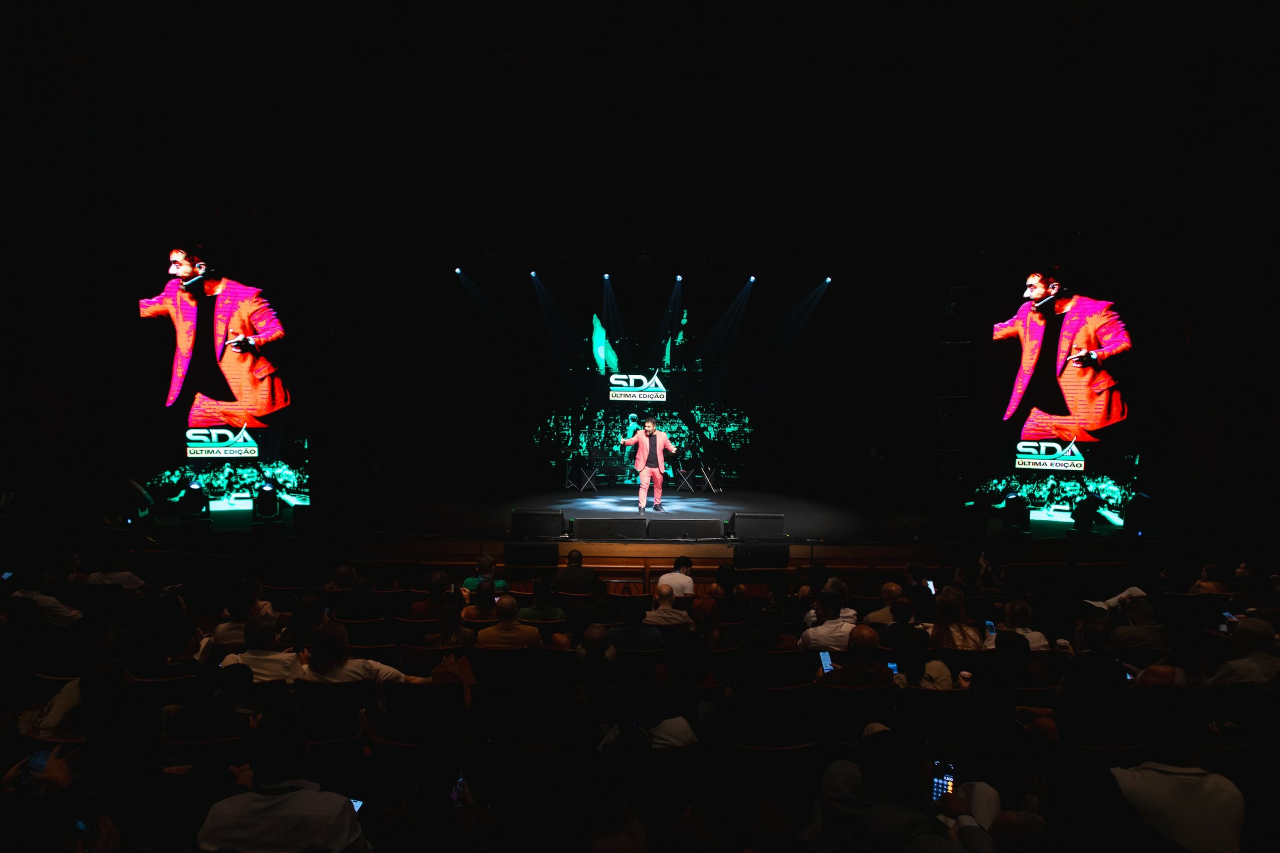 Stage with performer in center, two large screens on sides showing the same image, audience seated in front.